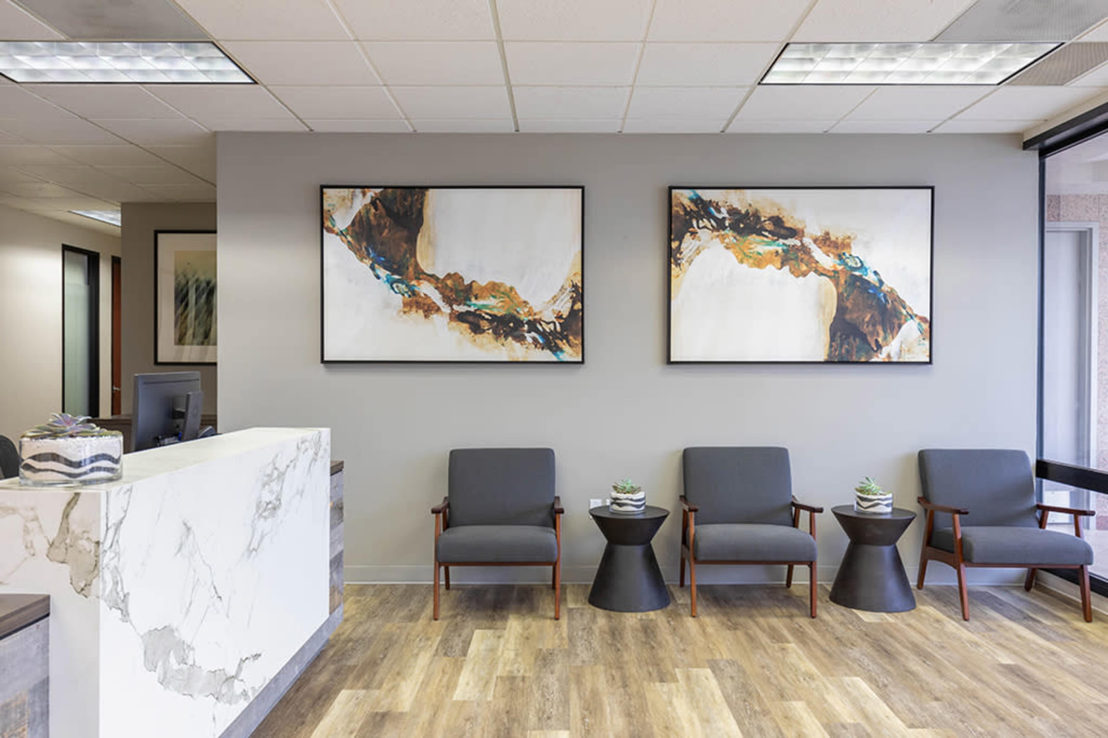 A modern waiting area featuring three gray chairs, small side tables, and two abstract paintings on the wall.