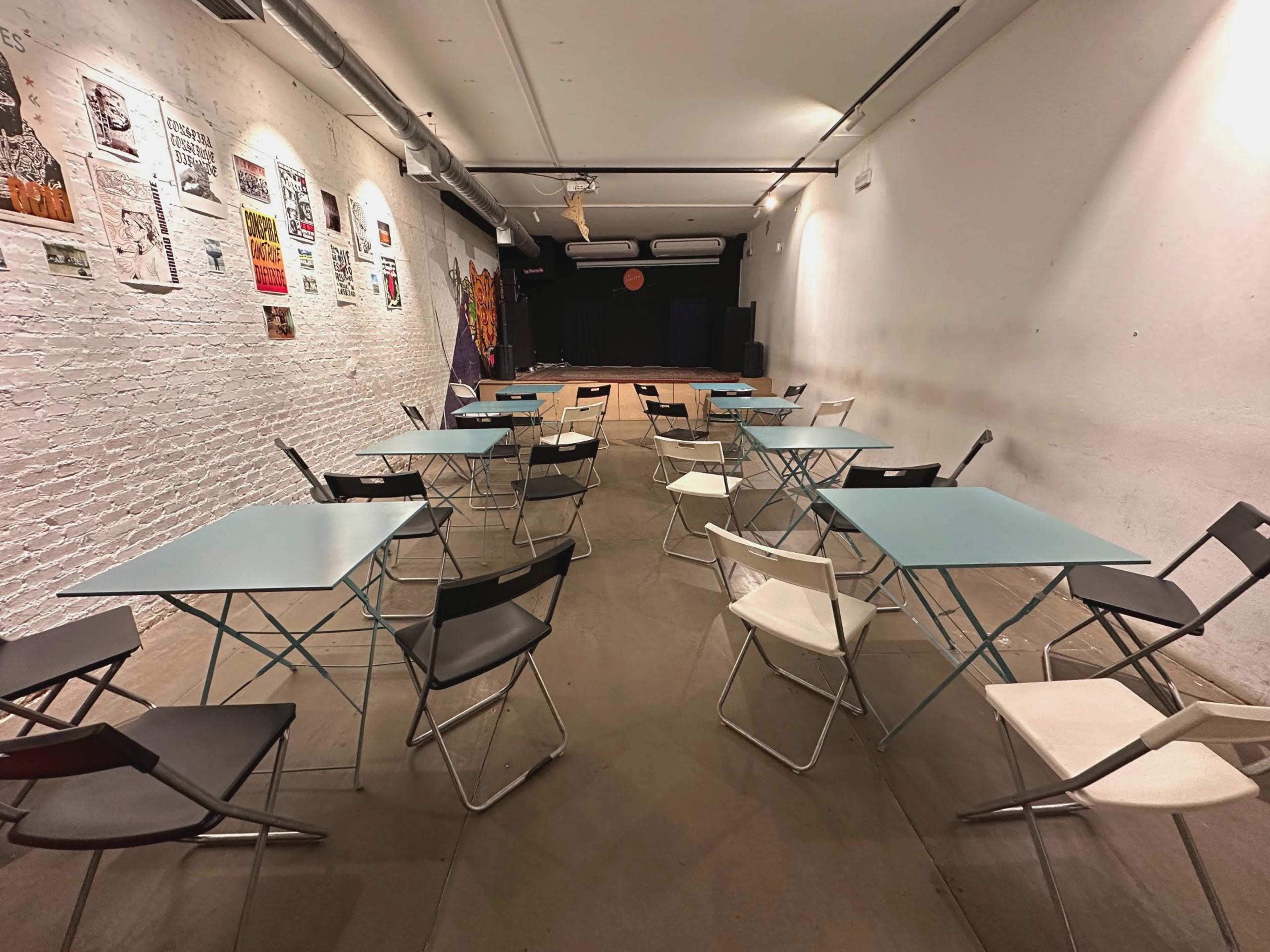 The image shows a dimly lit room arranged with multiple folding tables and chairs, facing a small stage against the far wall.