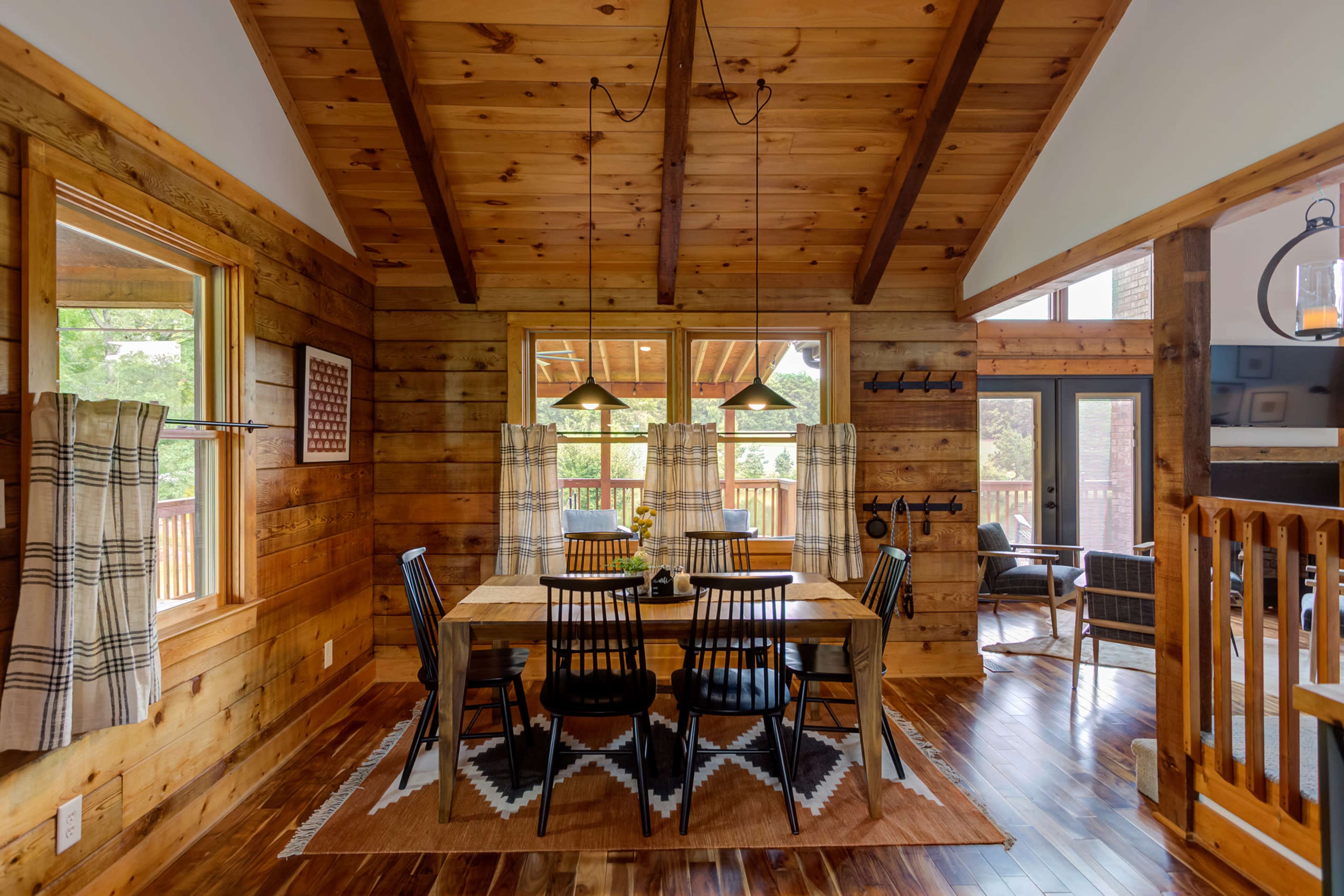 A wooden dining area features a table surrounded by black chairs, with large windows revealing an outdoor view and warm wooden decor throughout.