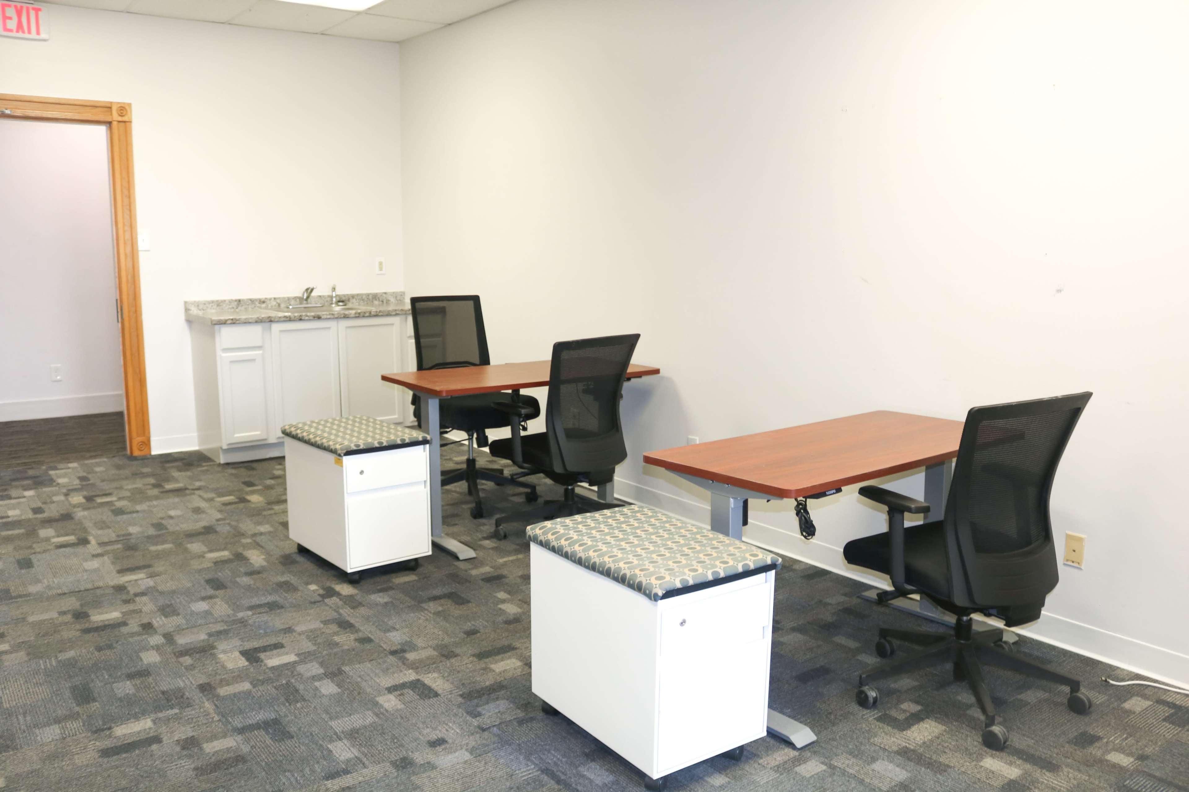 The image shows a small office space with two workstations, each featuring a desk, an ergonomic chair, and a storage unit, along with a kitchenette area in the background.