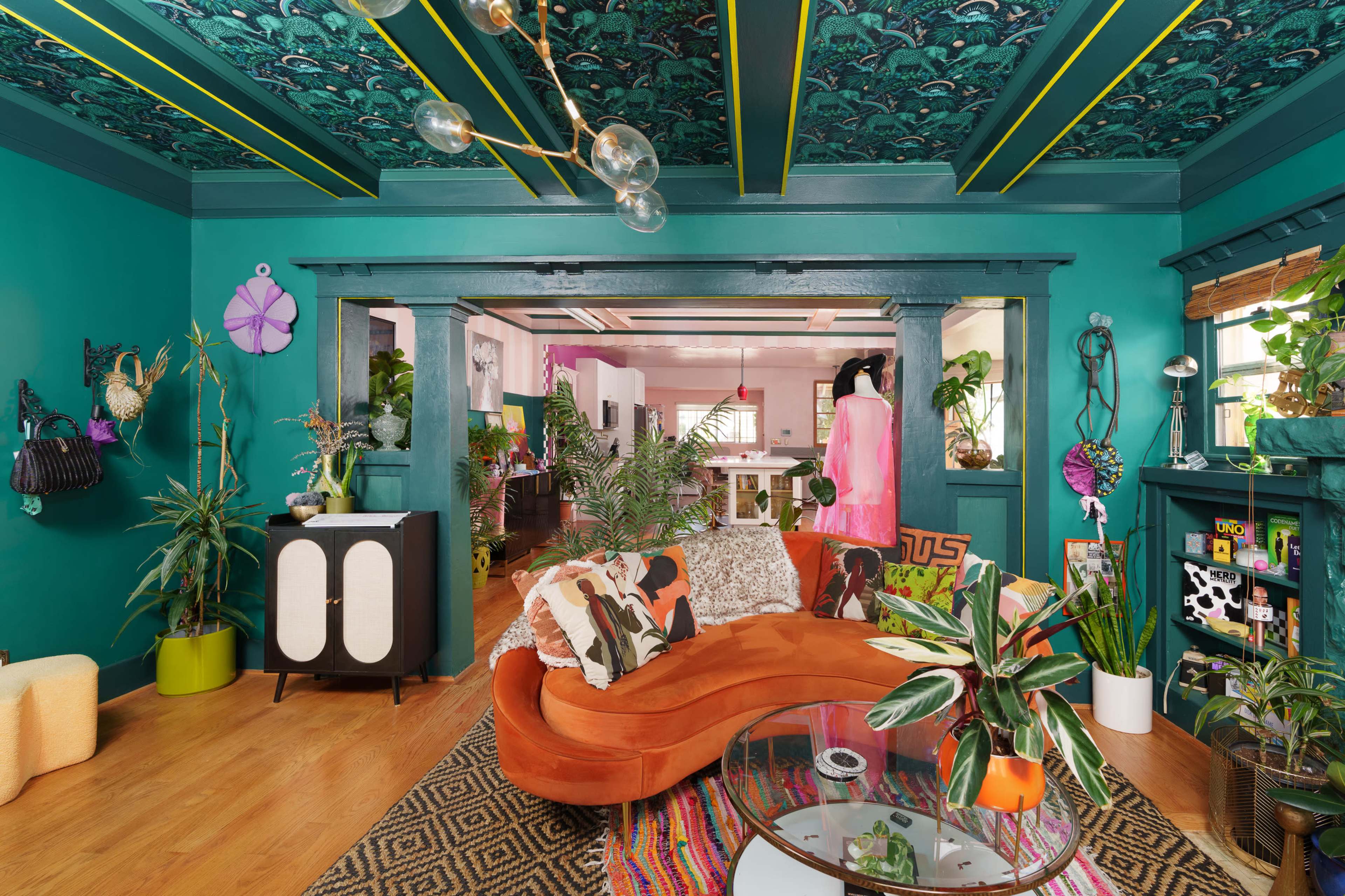 The image shows a vibrant living room with bold green walls, a curved orange sofa, plants, and decorative elements, creating an eclectic and lively atmosphere.
