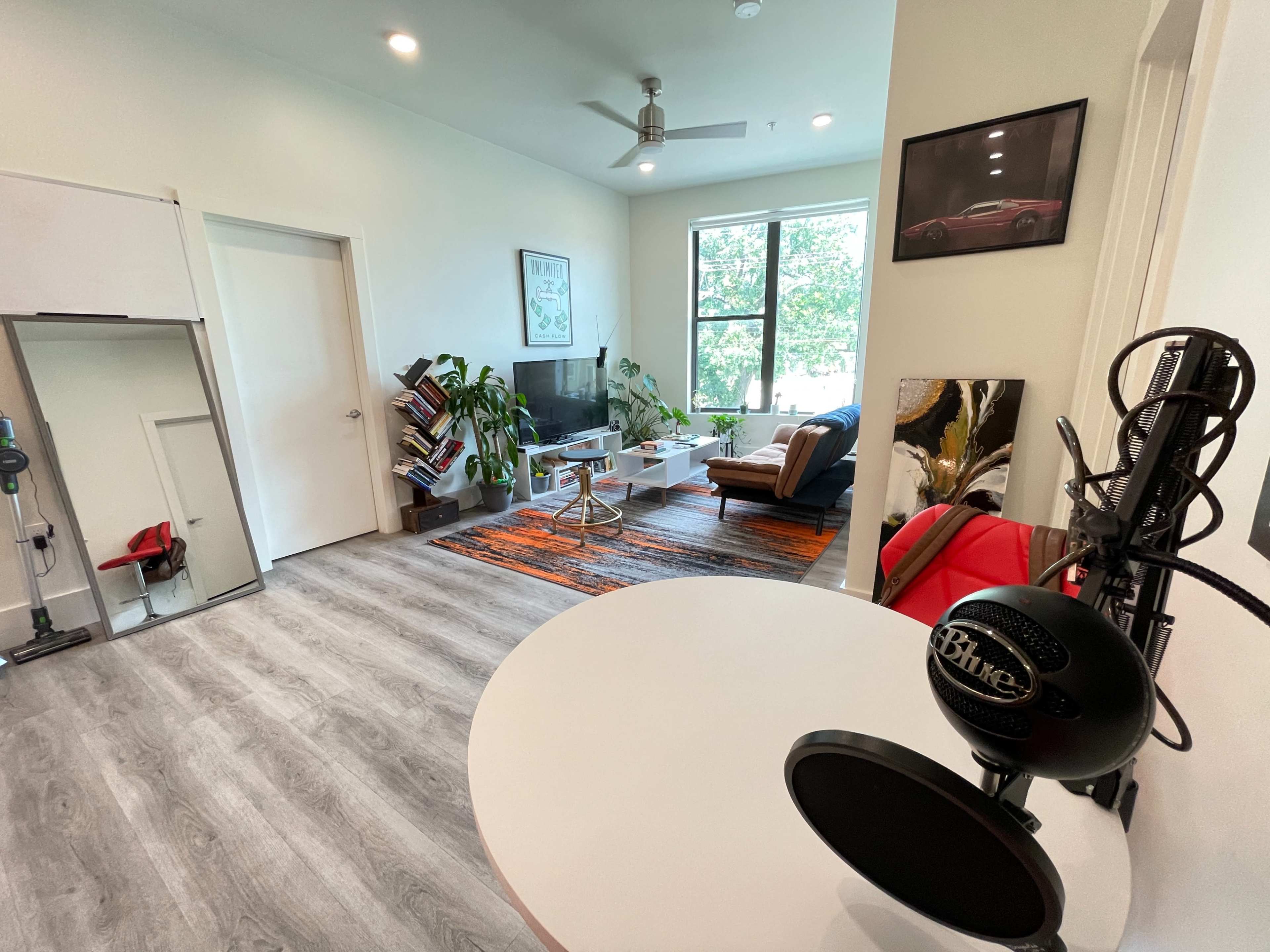 A bright living space featuring a white wall, a gray floor, a large window with natural light, a black entertainment unit, a brown armchair, and a rug with bold colors.