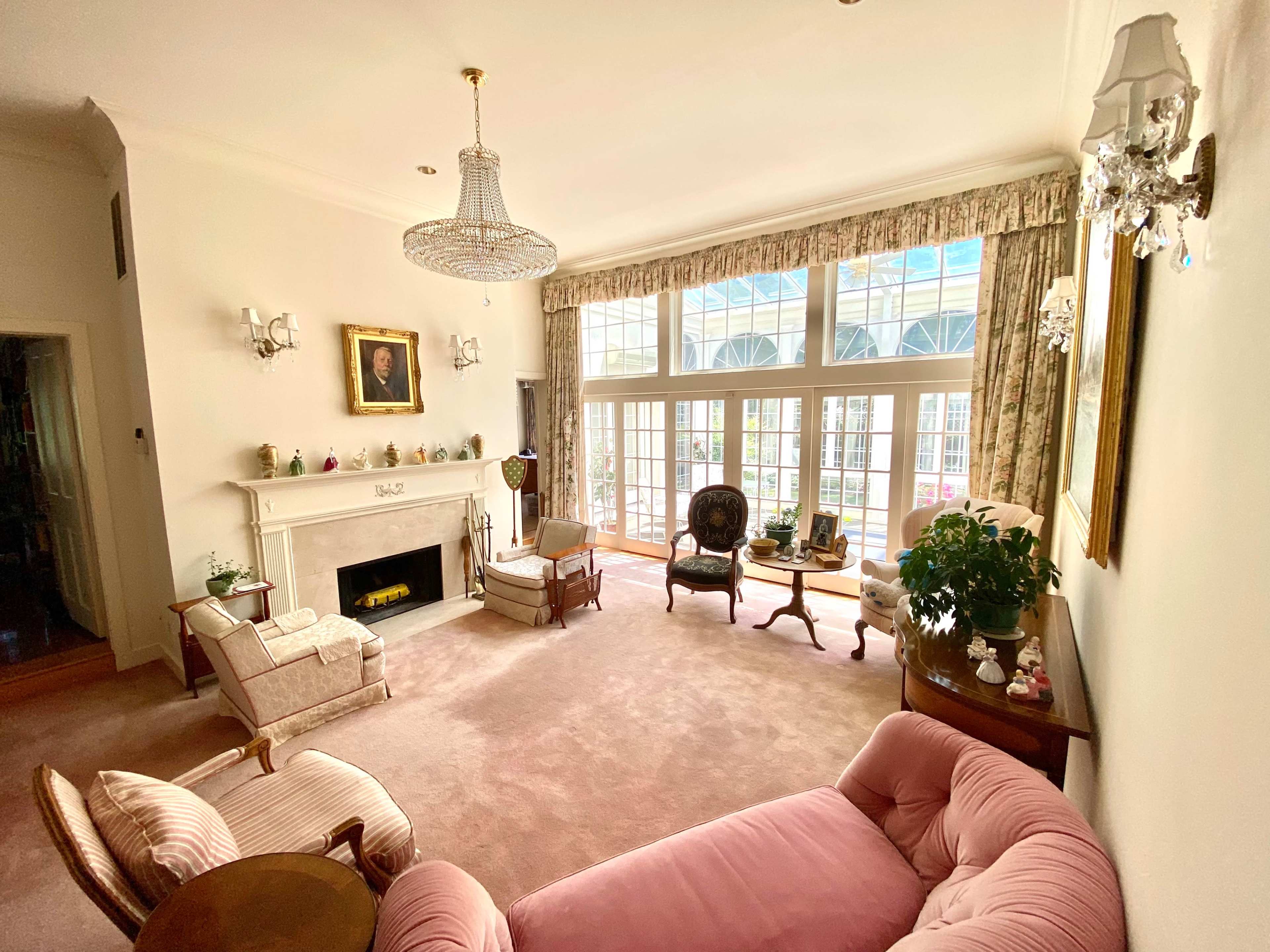 A spacious living room features large windows, light-colored walls, a pink sofa, elegant furniture, and a fireplace.