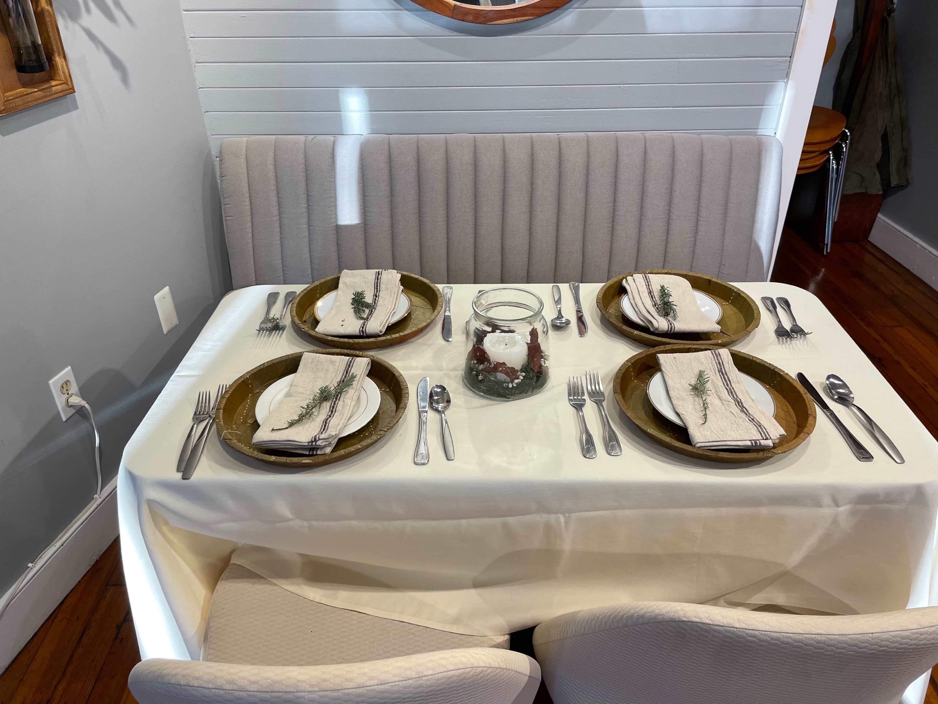 A neatly set dining table with four place settings, featuring plates, cutlery, and decorative napkins, is positioned beside a booth against a wall.