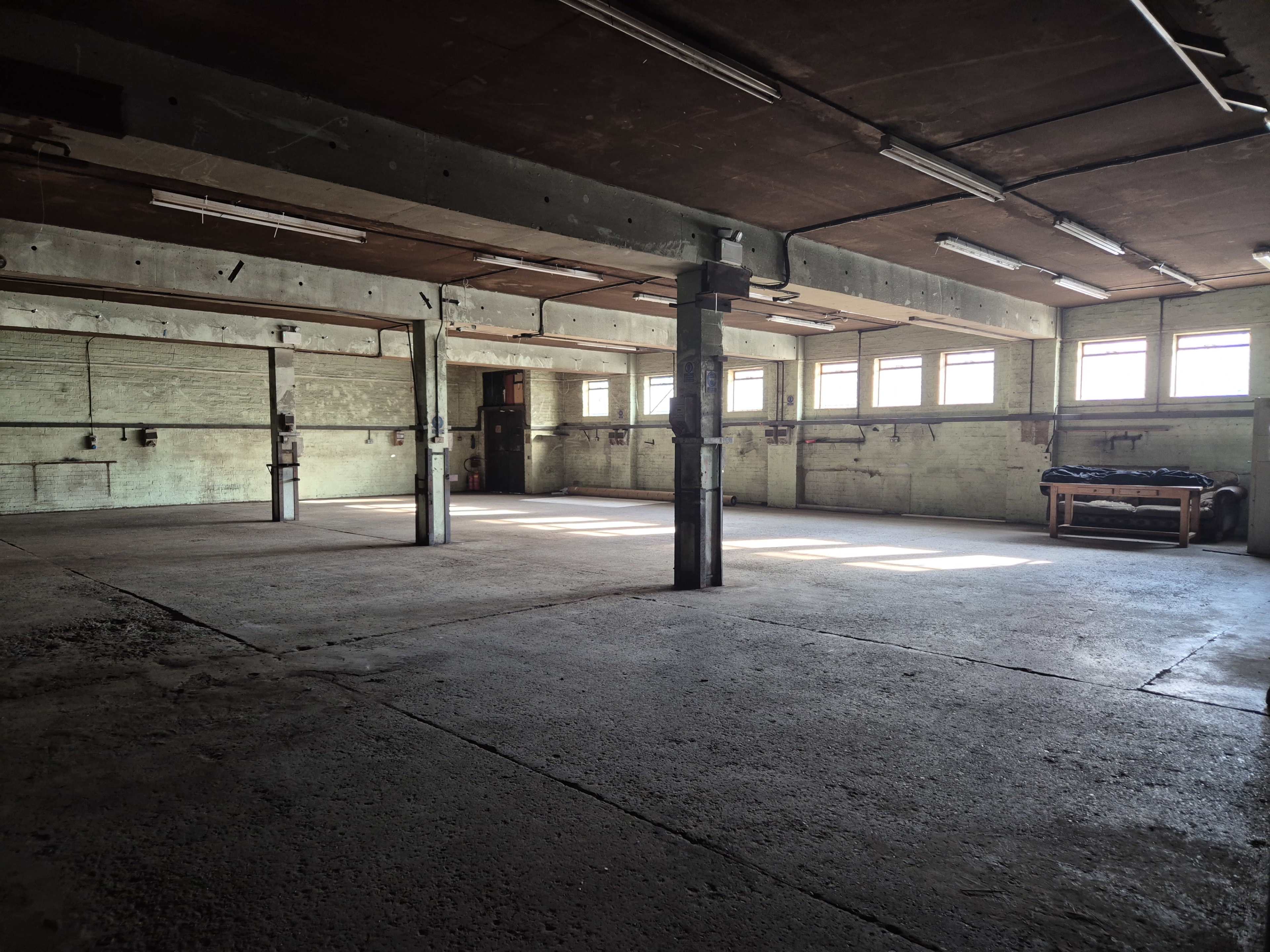 The image shows an empty, industrial-style room with concrete flooring, large windows, and exposed beams.