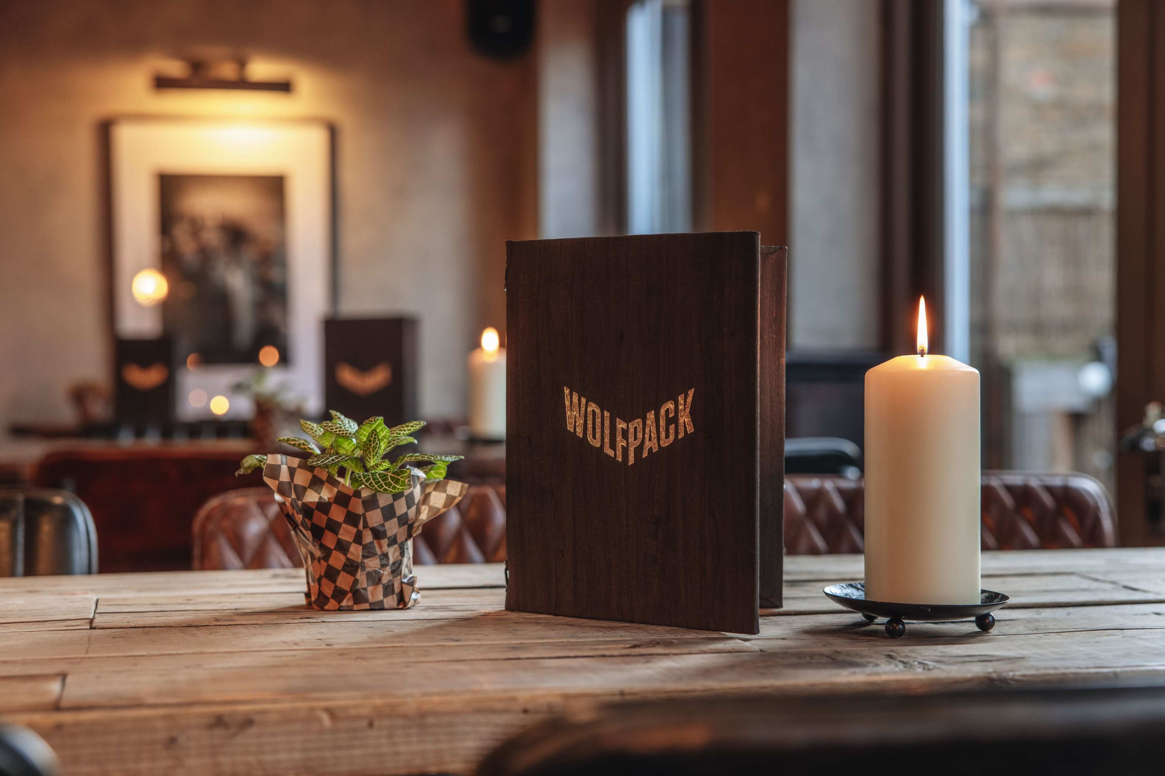 A wooden table in a restaurant features a decorative menu labeled "WOLFPACK," a potted plant, and a lit candle.