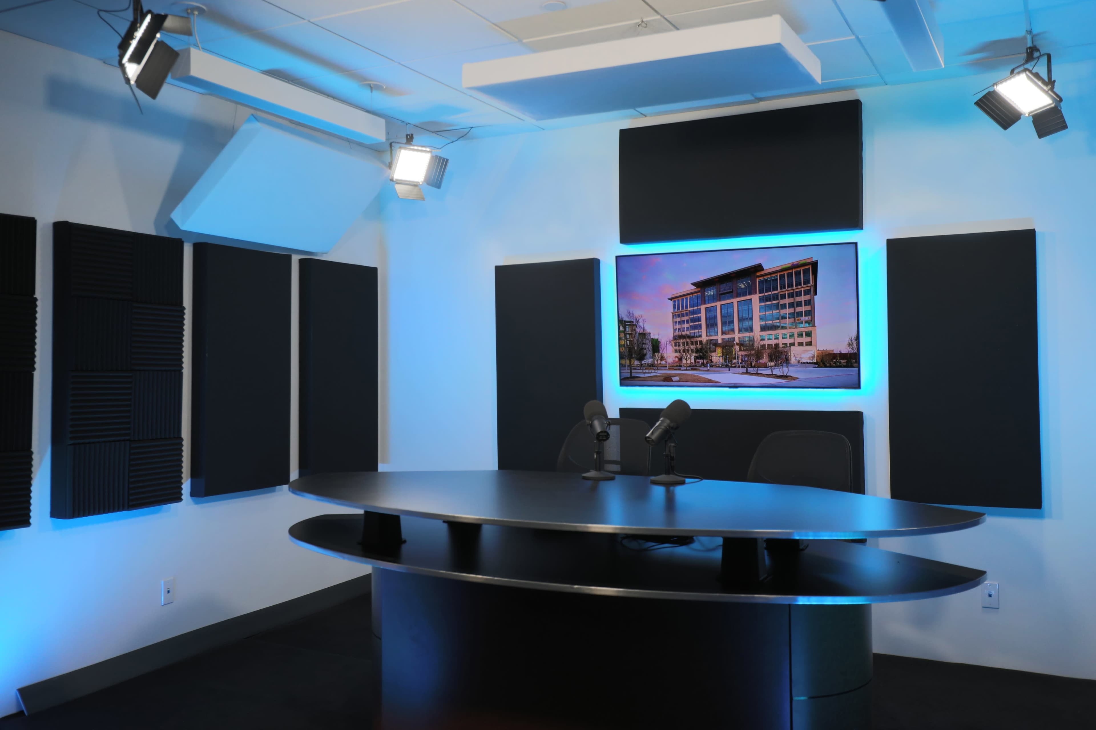 A modern broadcast studio with a black curved table, two microphones, and an illuminated screen displaying a building.