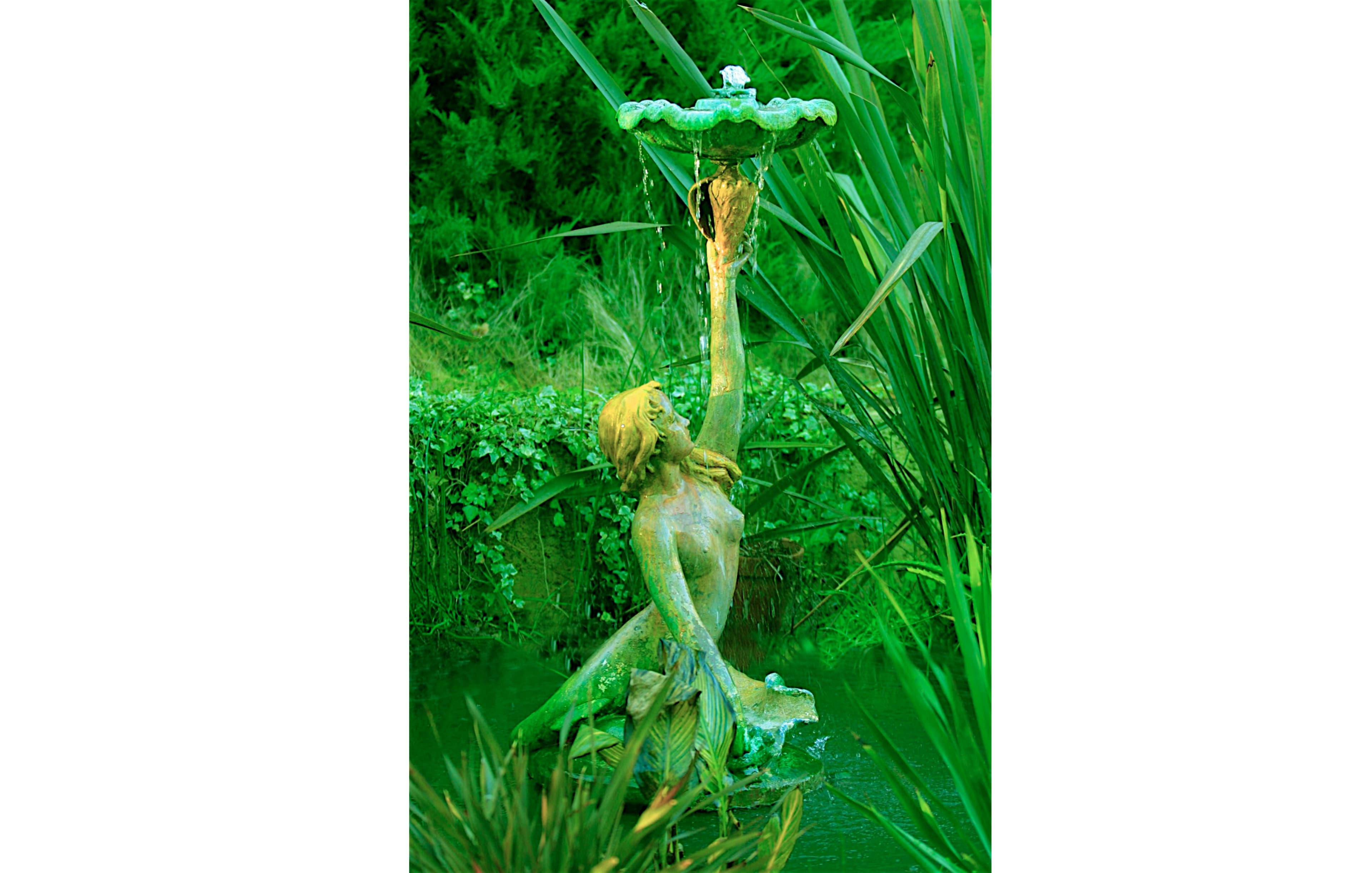 A green-tinted fountain sculpture of a woman holding a basin is situated by a pond surrounded by lush vegetation.