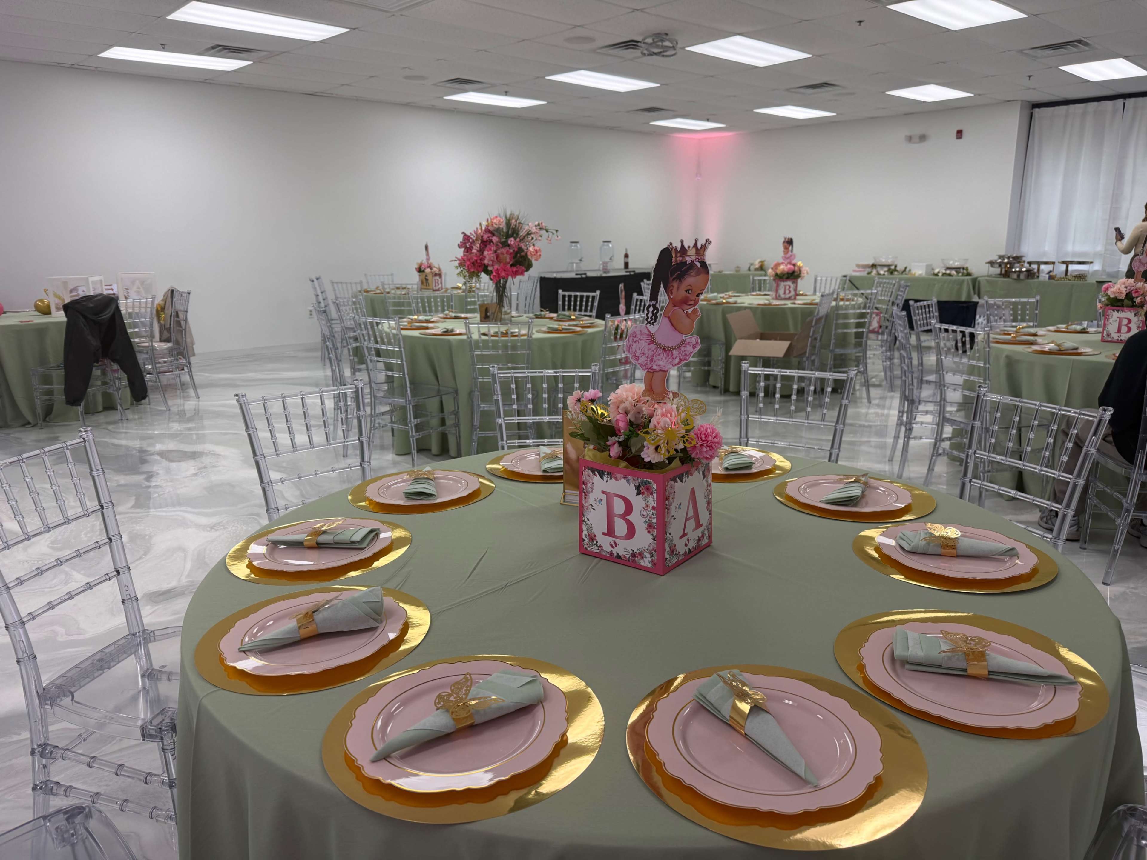A banquet hall is set up for an event, featuring circular tables with pale green linens, gold chargers, and pink plates, complemented by floral centerpieces and decorative elements.