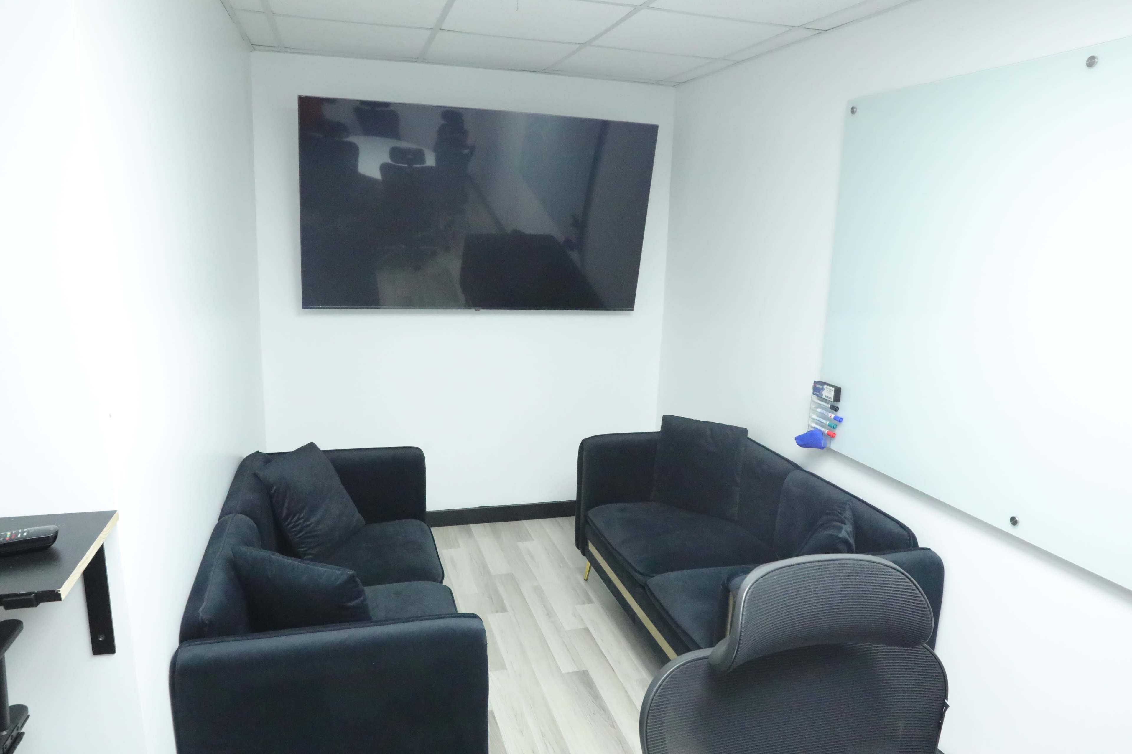 A small meeting room with two black sofas facing a large wall-mounted TV and a whiteboard.