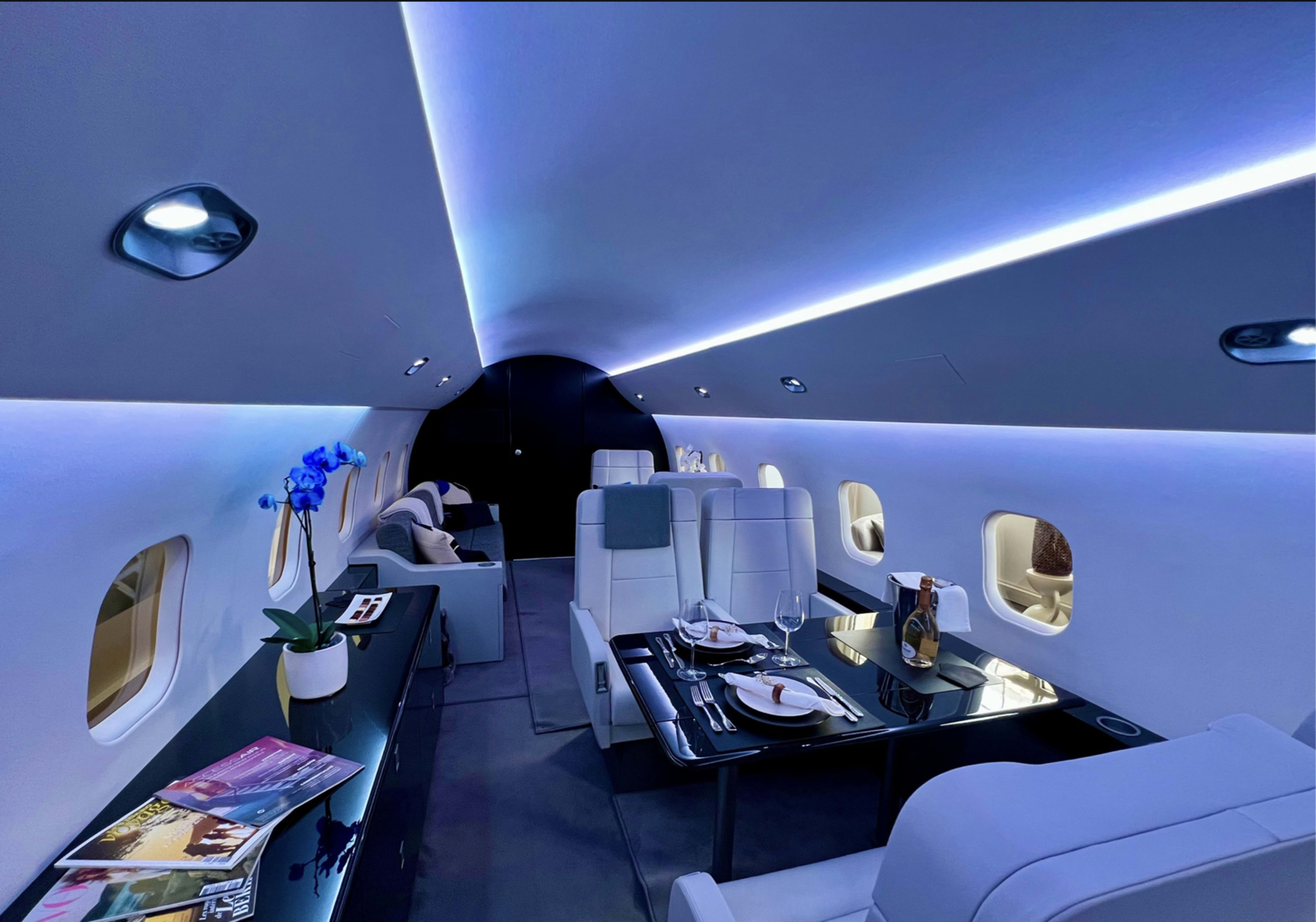 The interior of a luxury private jet features sleek white seating, a polished dining table set with tableware, and ambient blue lighting.
