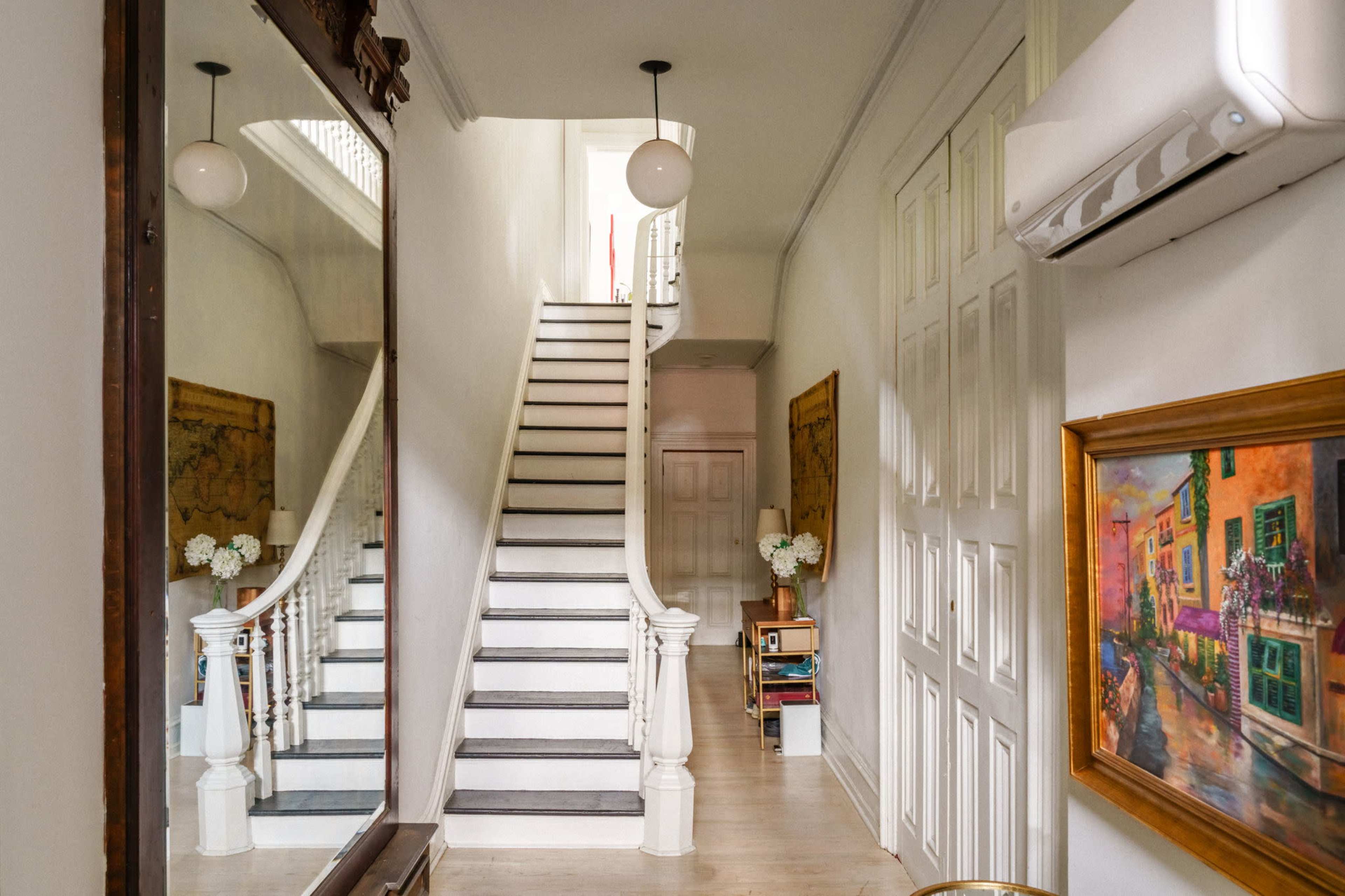 The image shows a well-lit hallway with a staircase leading to an upper floor, flanked by white walls and decorated with artwork and a large mirror.