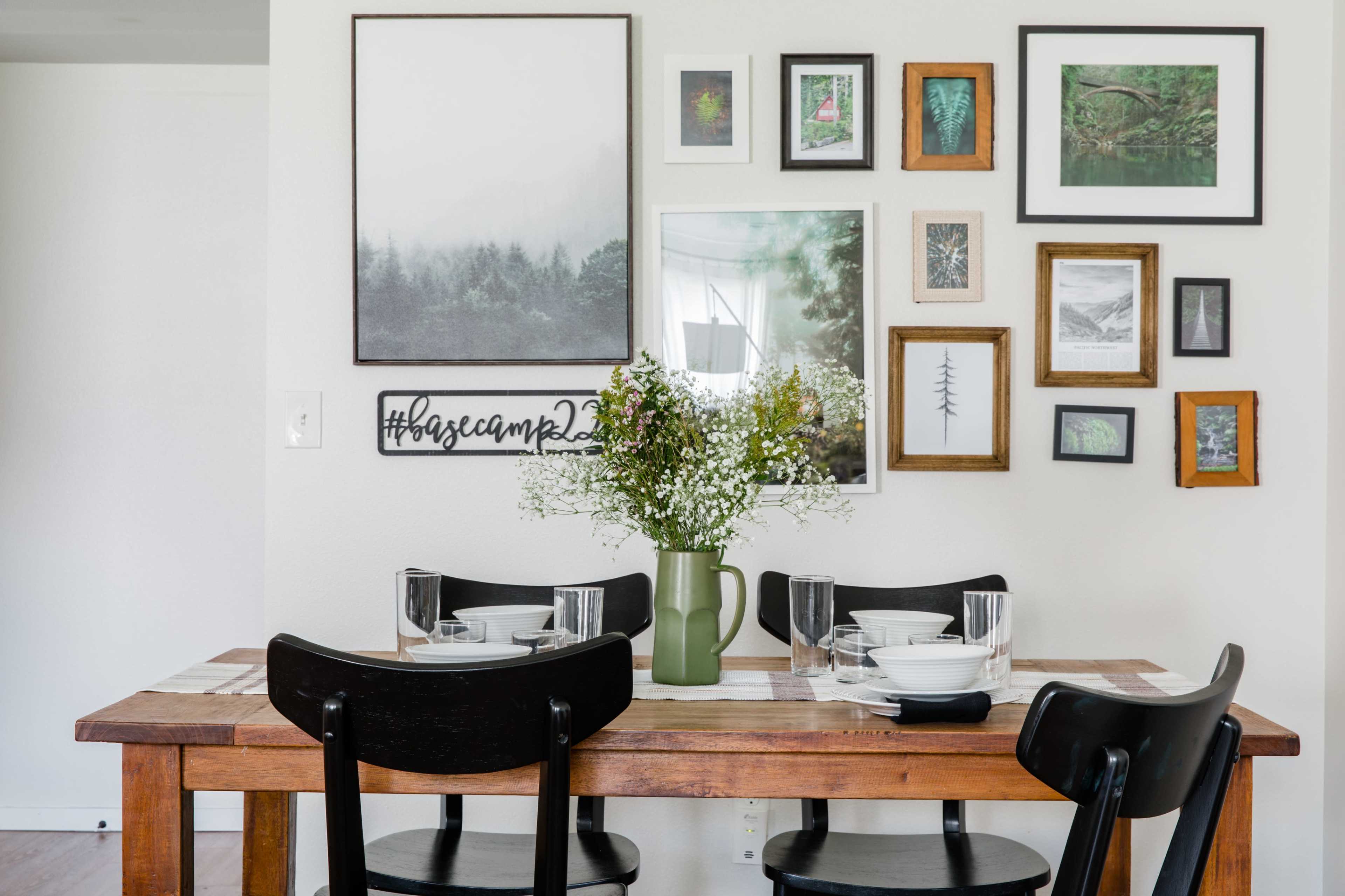 A wooden dining table is set with plates and glasses, surrounded by black chairs, and decorated with a wall of framed nature-themed artwork.