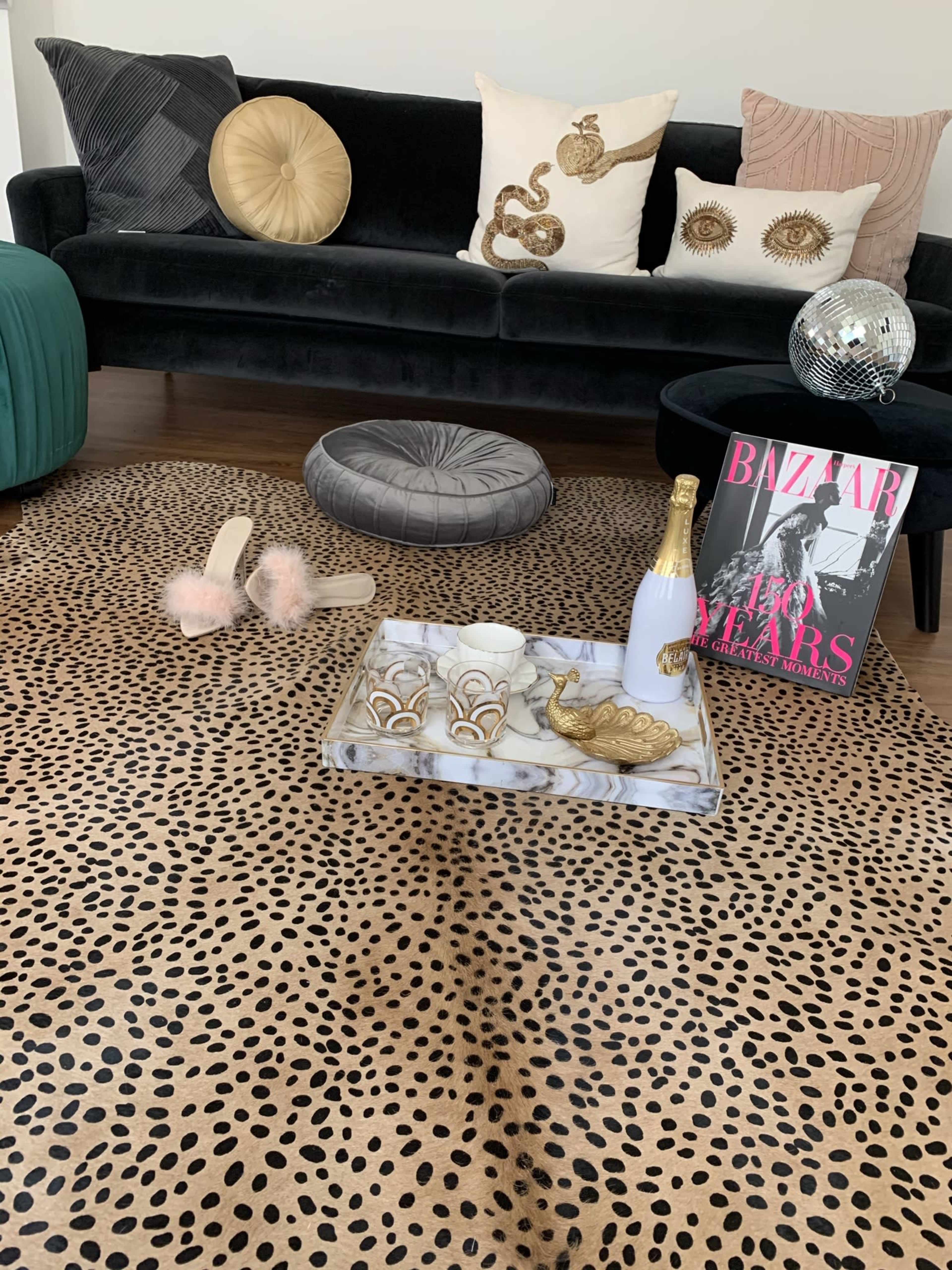 A stylish living room features a black velvet sofa with decorative pillows, a leopard print rug, a tray with decorative items, a magazine, and a pair of slippers nearby.