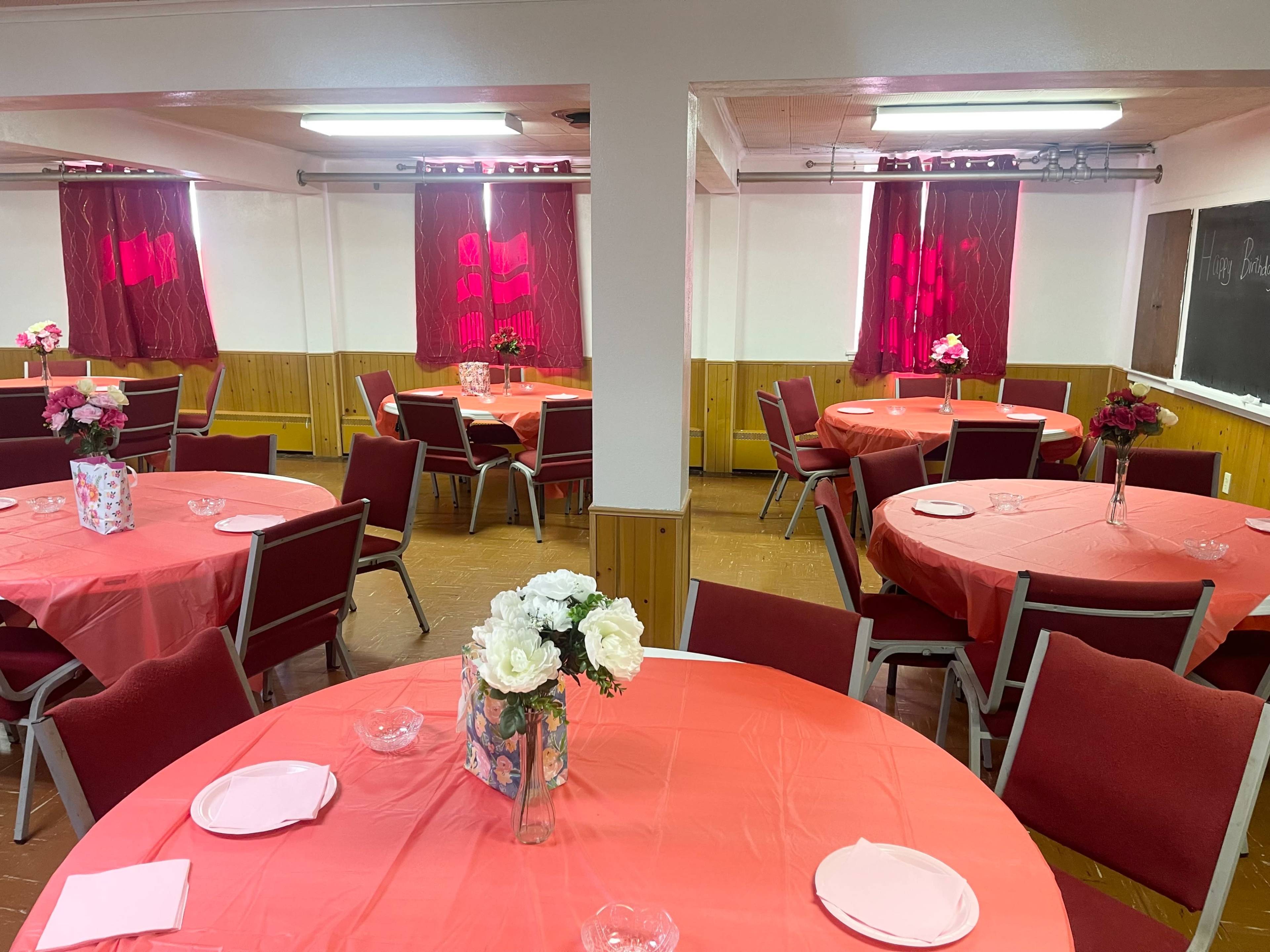 The image shows a banquet hall with round tables covered in orange tablecloths, each set with plates and flower centerpieces, and red curtains framing the windows.
