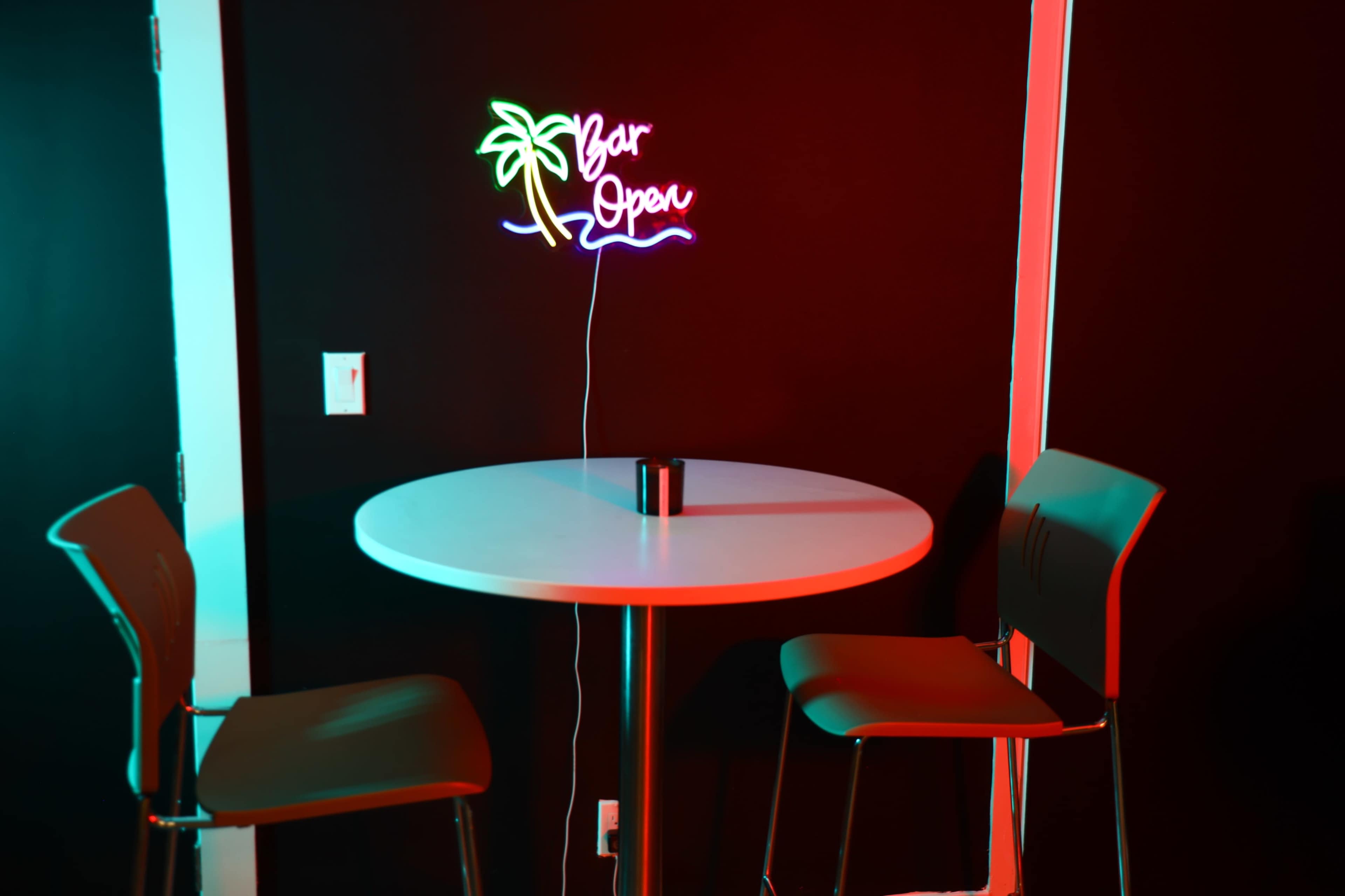 A round table with two chairs is positioned under a neon sign that says "Bar Open," set against a dark wall.