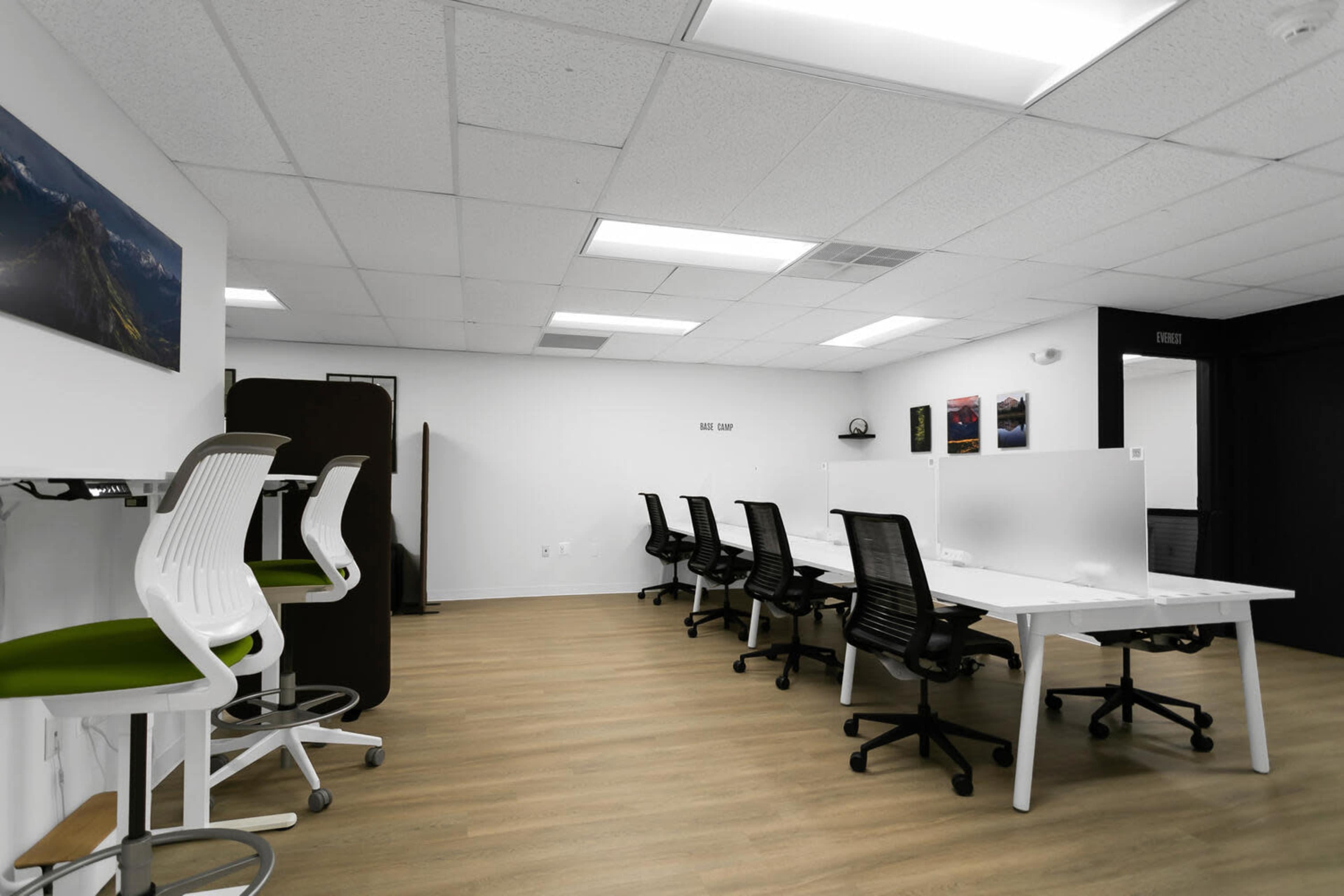 The image shows a modern office space featuring several workstations with ergonomic chairs, a white dividing panel, and minimal decorative elements on the walls.