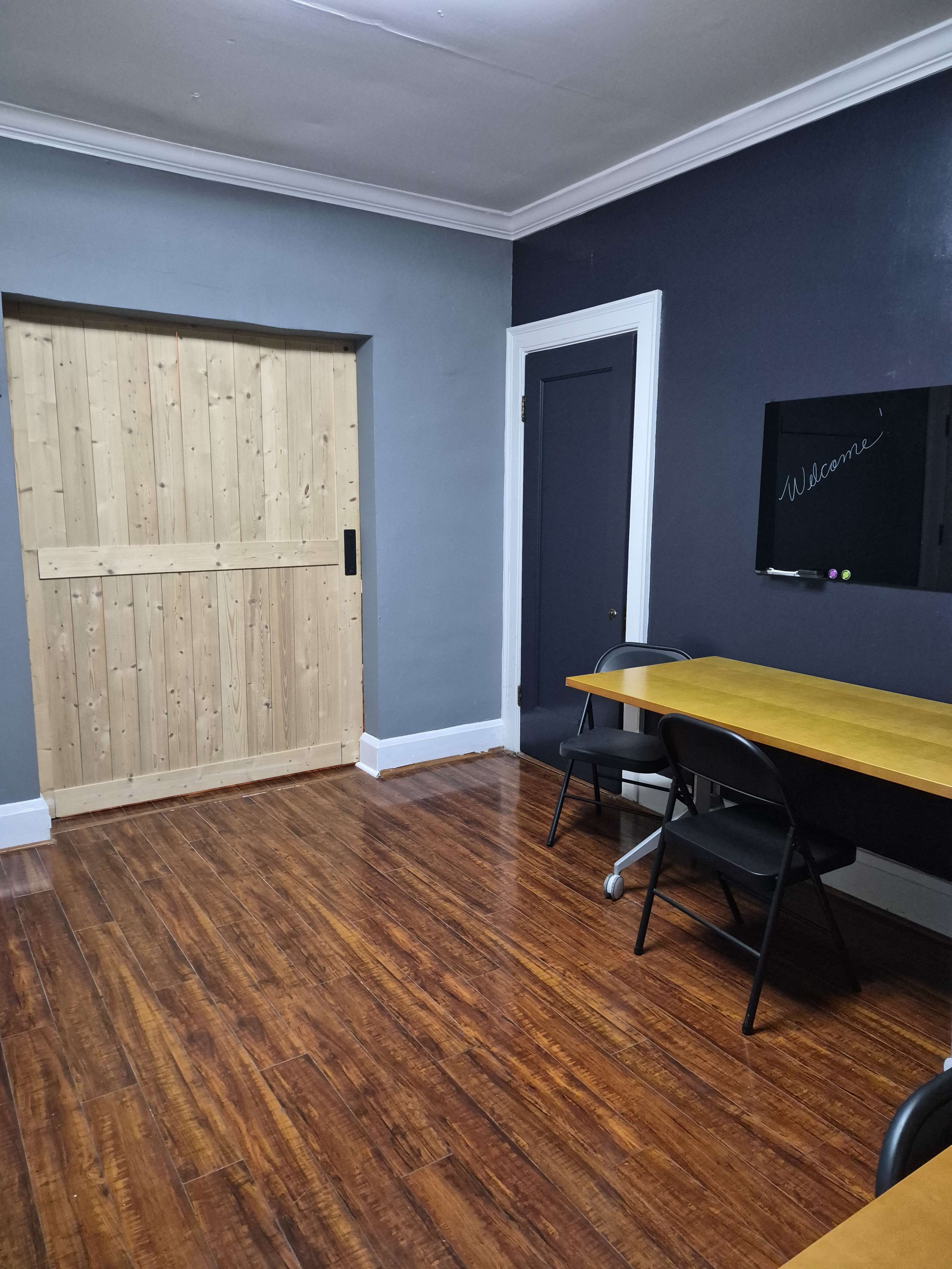The image shows a simple office space featuring a wooden sliding door, a chalkboard on the wall, and two desks with black chairs on a polished wooden floor.
