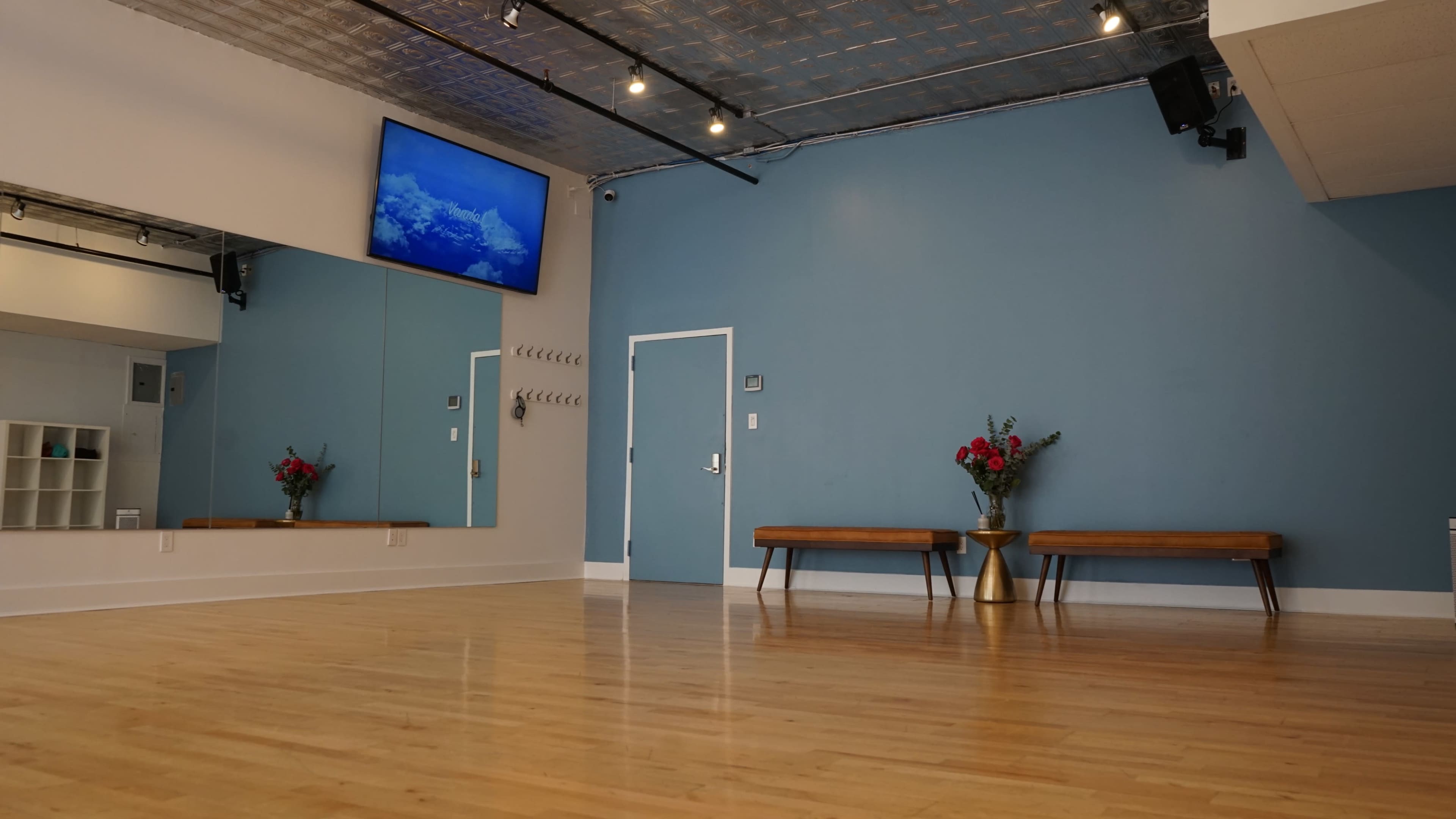 The image shows a spacious room with wooden flooring, a television mounted on the wall, a set of mirrors, two benches, and a vase of flowers on a table against a blue wall.