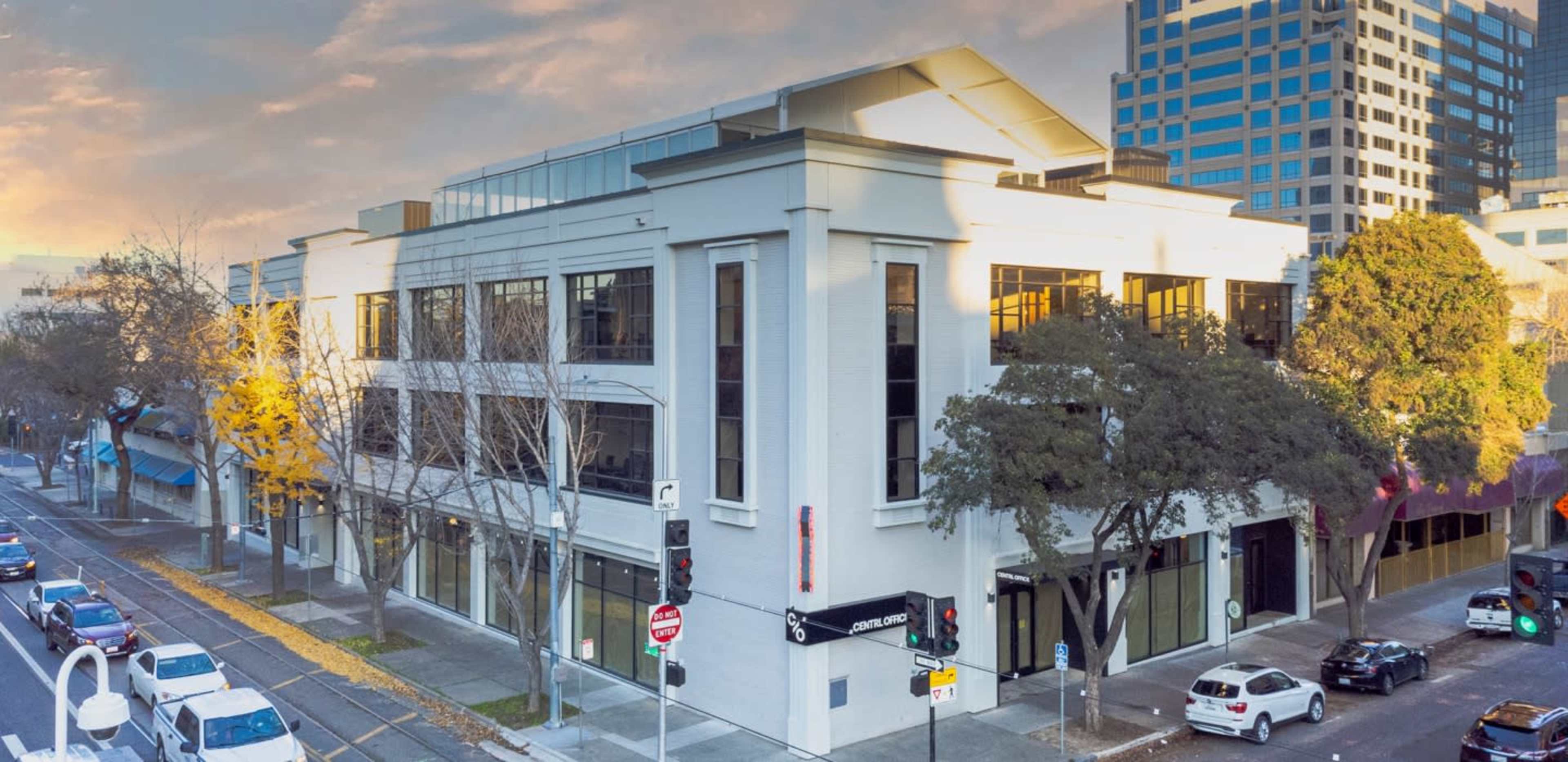 A modern multi-story building located on a city street, with large windows and surrounding trees, under a partly cloudy sky.