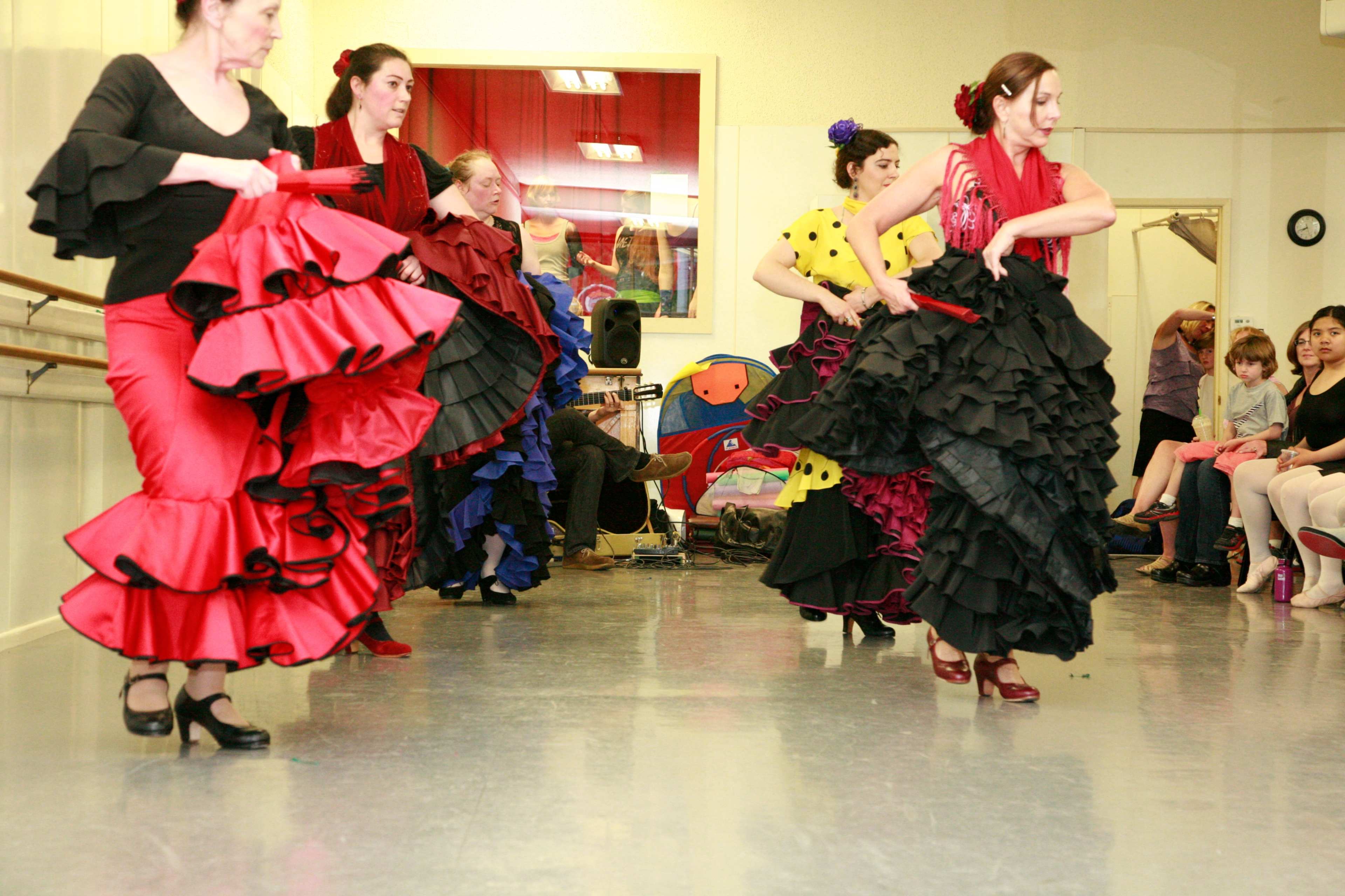 A group of women in vibrant, ruffled dresses practices a dance routine in a studio with a mirror and wooden barre.