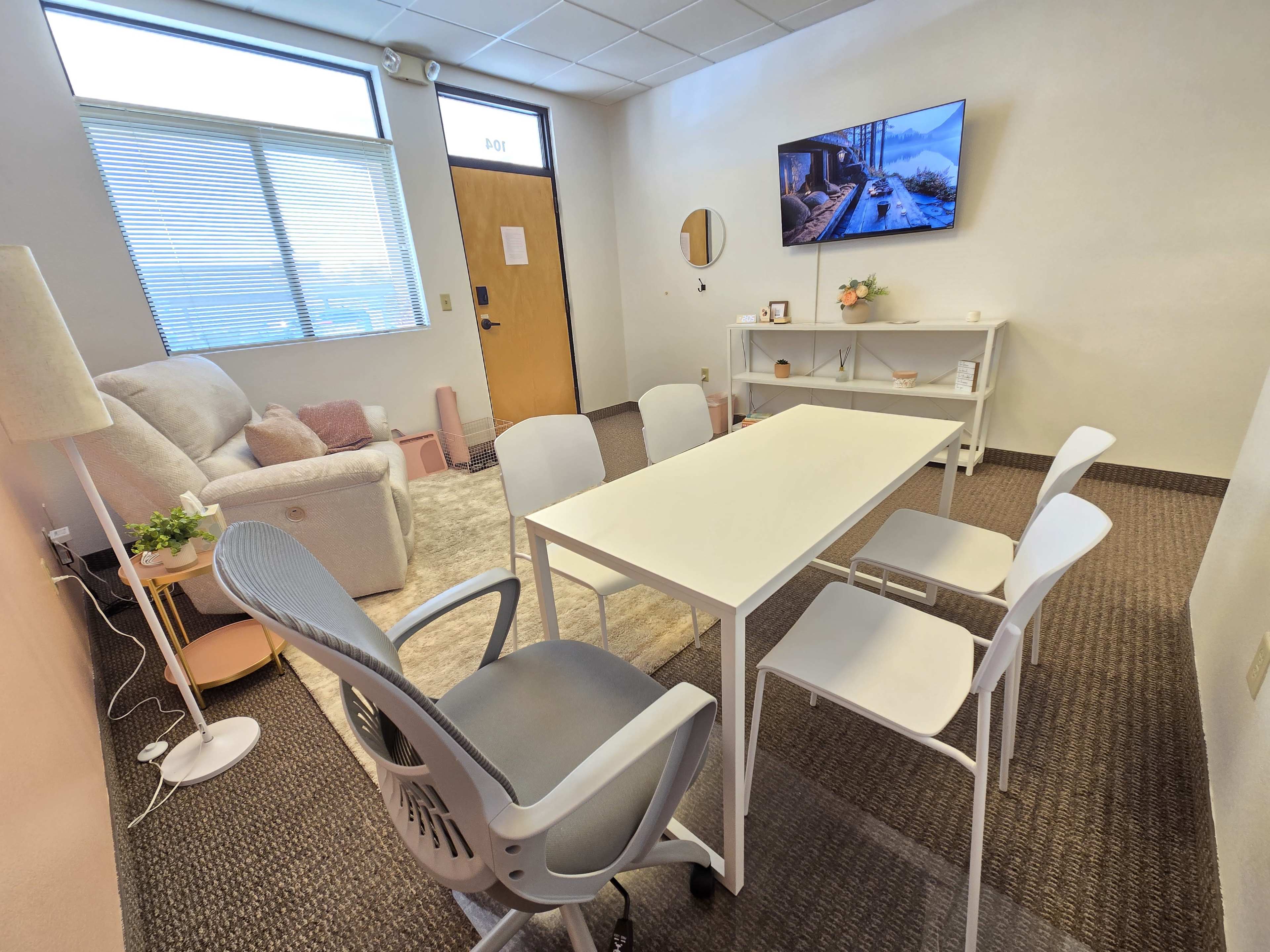 The image shows a small, light-colored office or meeting room with a table surrounded by four chairs, a sofa in the corner, a lamp, and a television mounted on the wall.