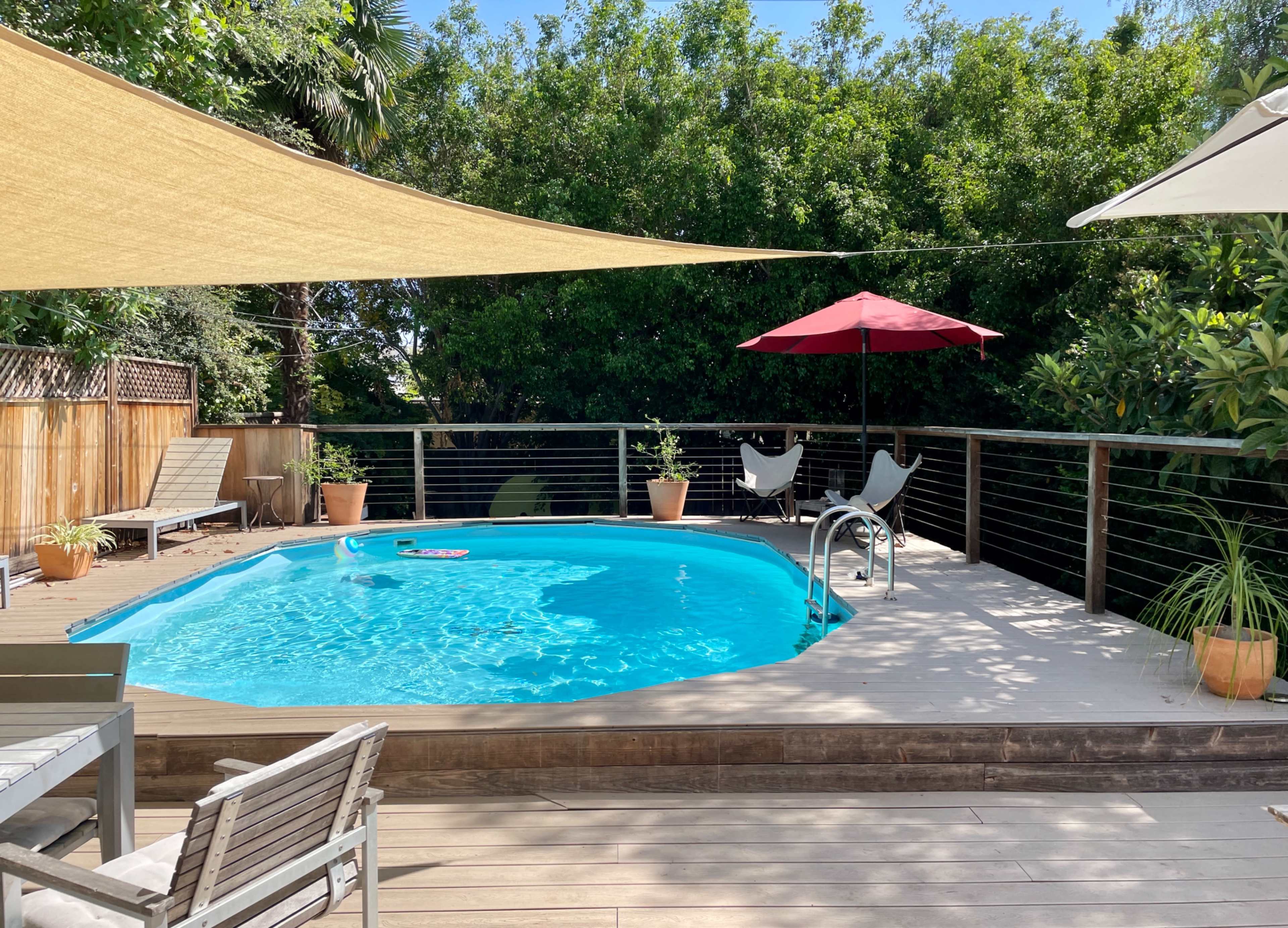 A backyard pool area features a clear blue pool surrounded by wooden decking, sunshades, potted plants, and outdoor furniture.
