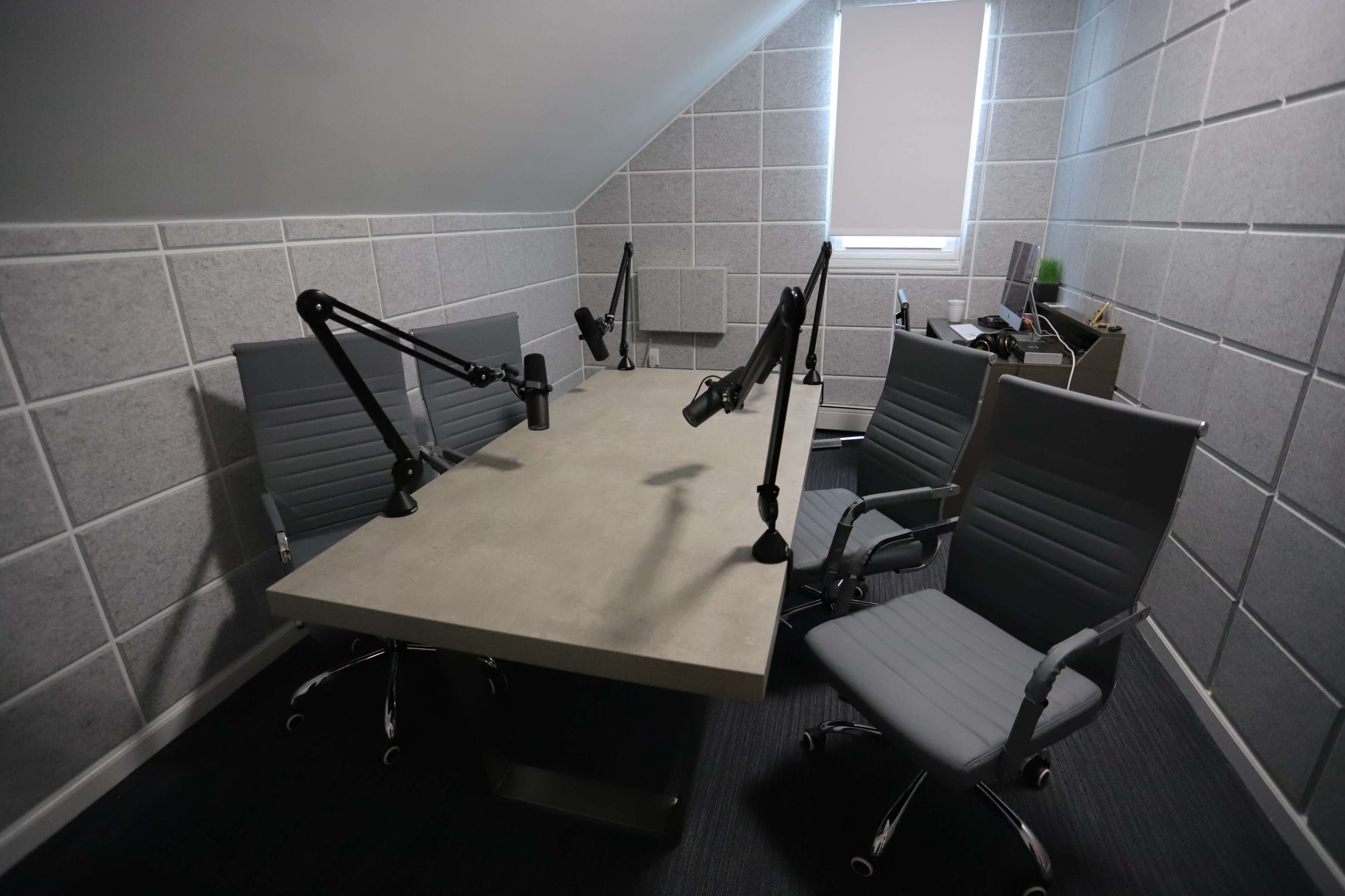 A small recording studio features a large table with microphones and three chairs arranged around it, all set against grey acoustic paneling.