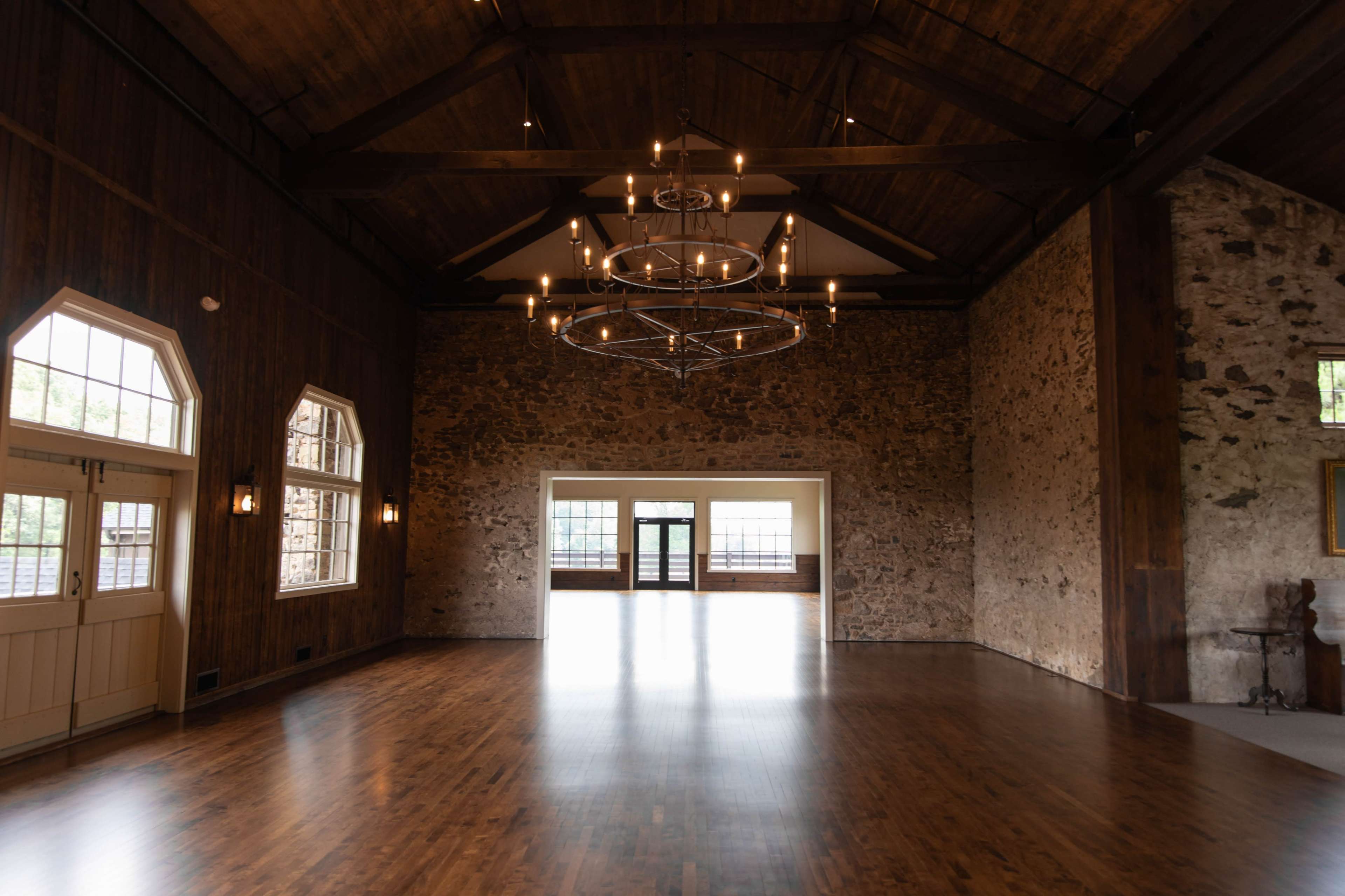 A spacious room with wooden beams, a stone wall, and large windows, illuminated by circular chandeliers.