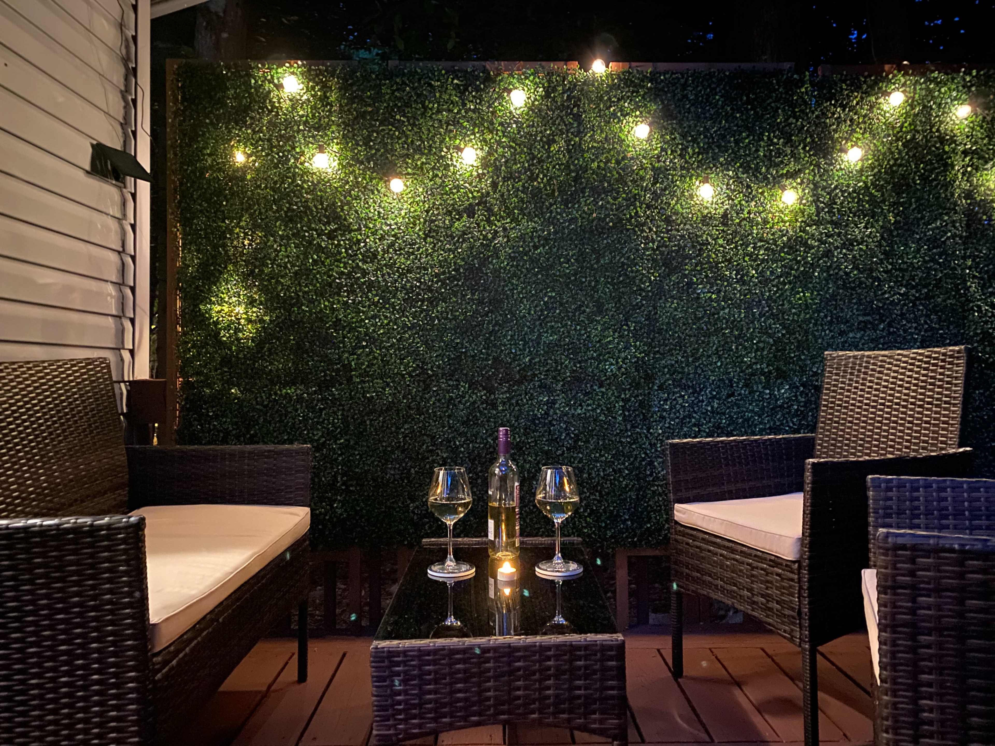 A cozy outdoor seating area with two wicker chairs, a glass coffee table, a bottle of wine, and lit string lights against a green hedge backdrop.