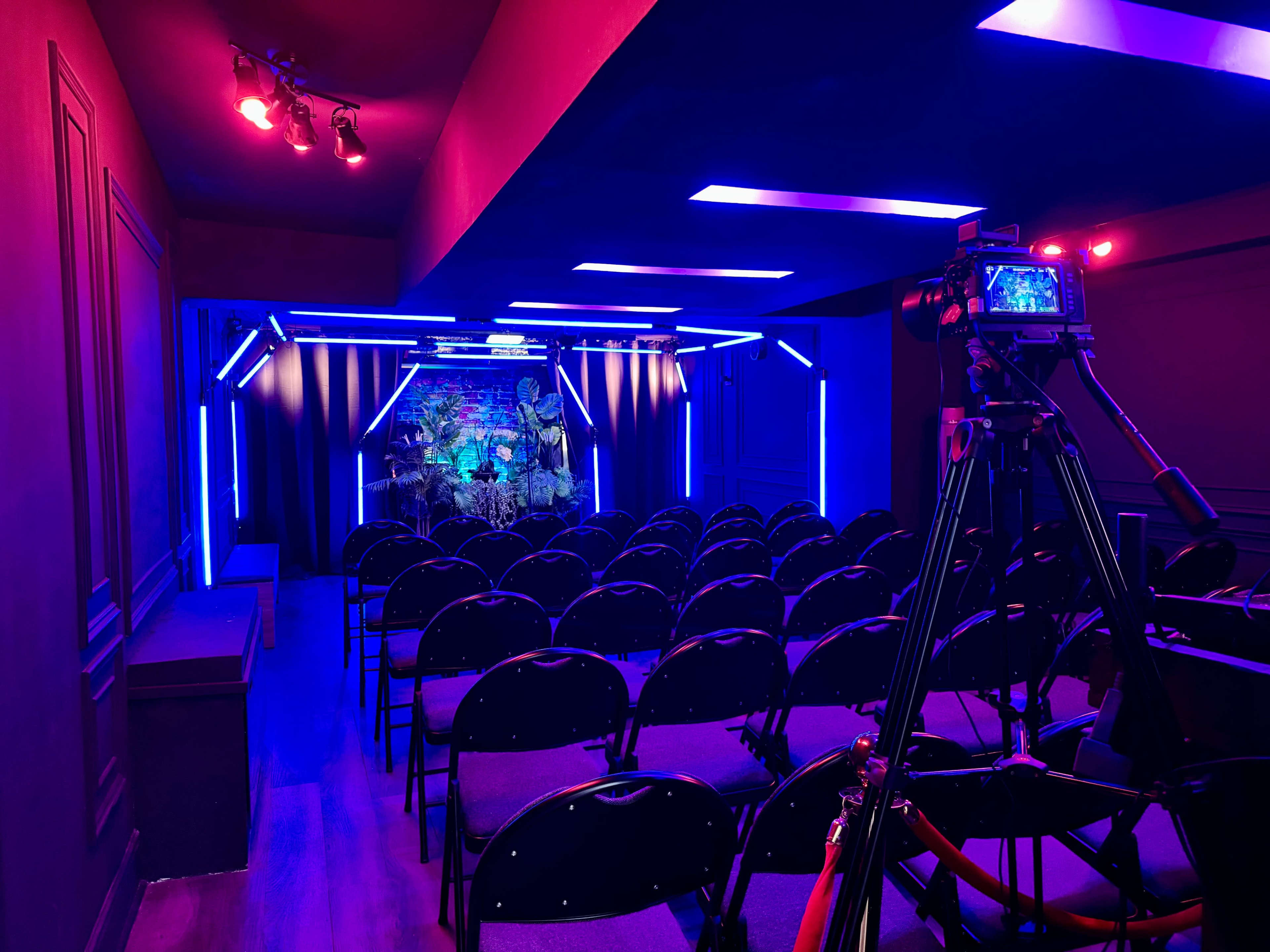 The image shows a small auditorium with rows of black chairs facing a brightly-lit stage decorated with colorful lighting and a backdrop.