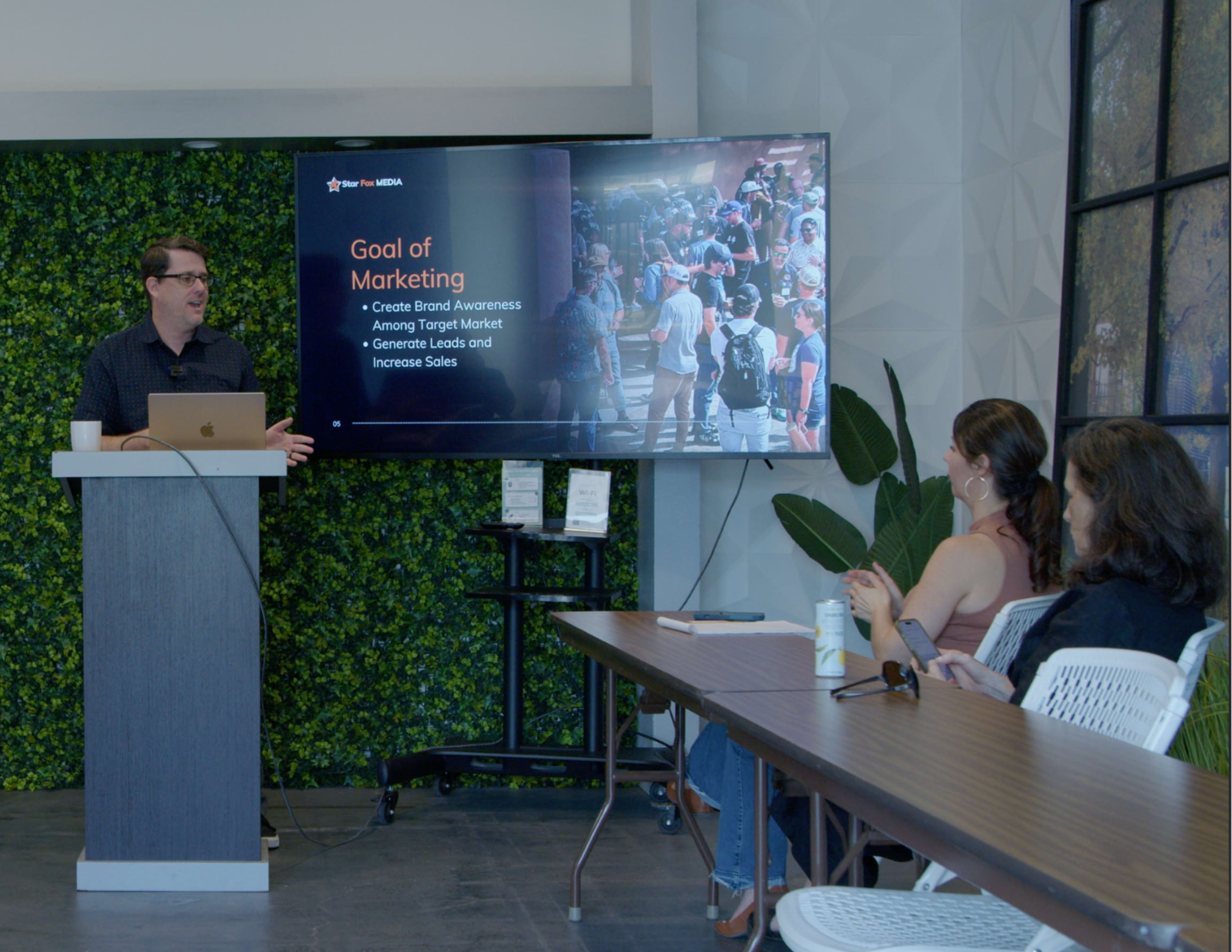 A presenter stands at a podium in front of a screen displaying marketing goals, while an audience sits at a long table attentively listening.