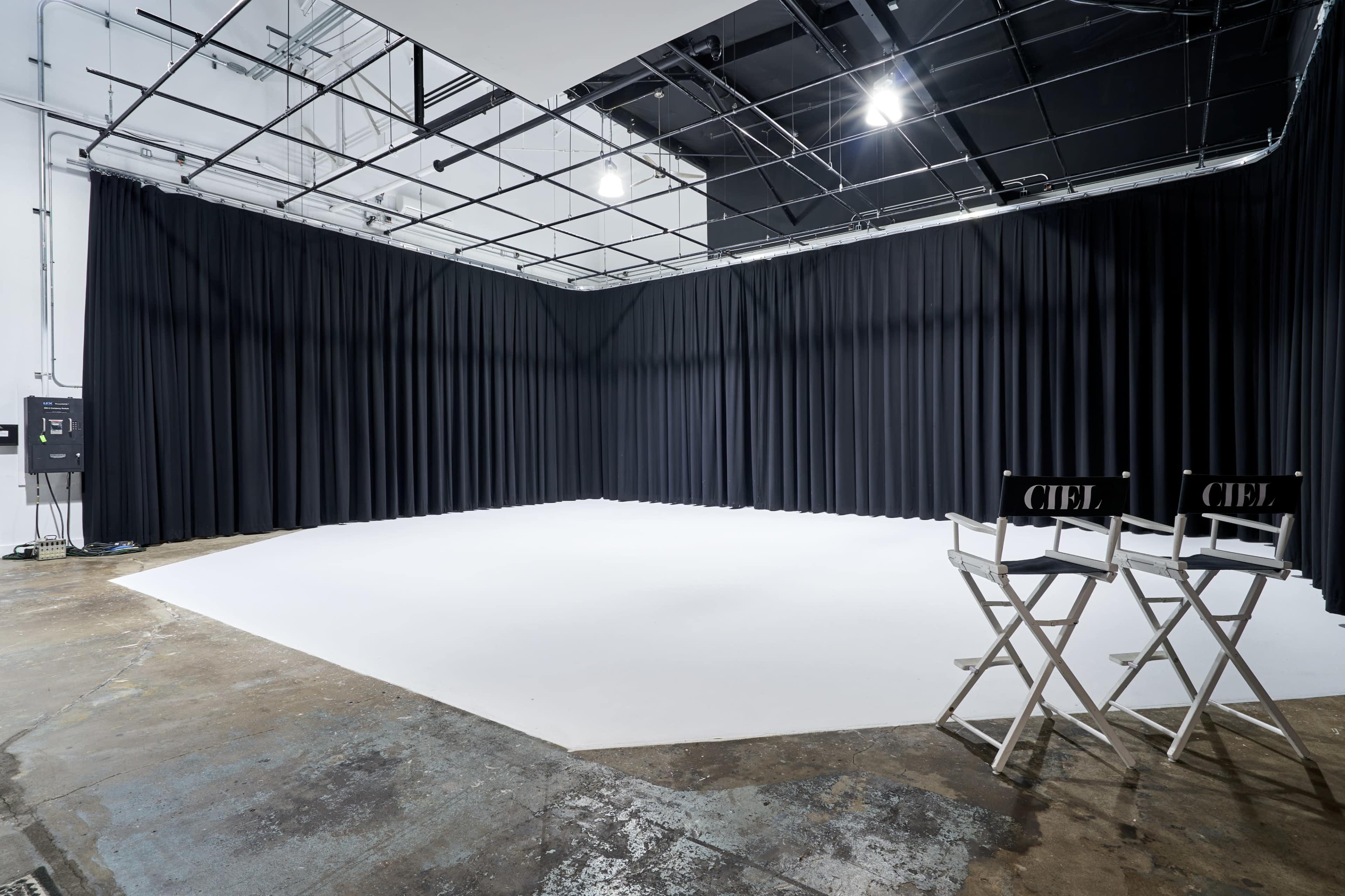 A spacious, empty studio with black curtains lining the walls and two director's chairs positioned on a white floor.