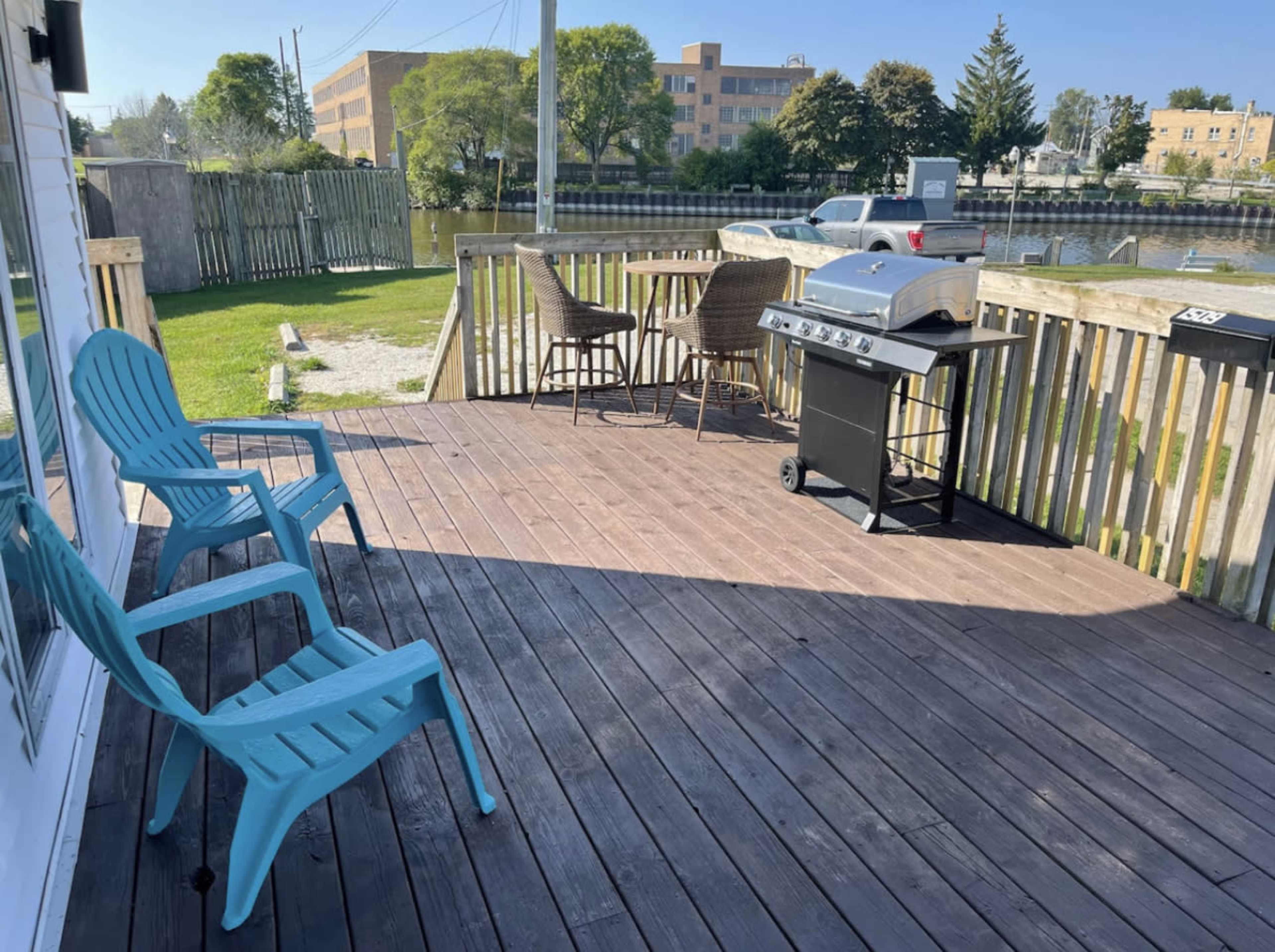 A wooden deck with blue chairs, a grill, and a view of a grassy area and buildings in the background.