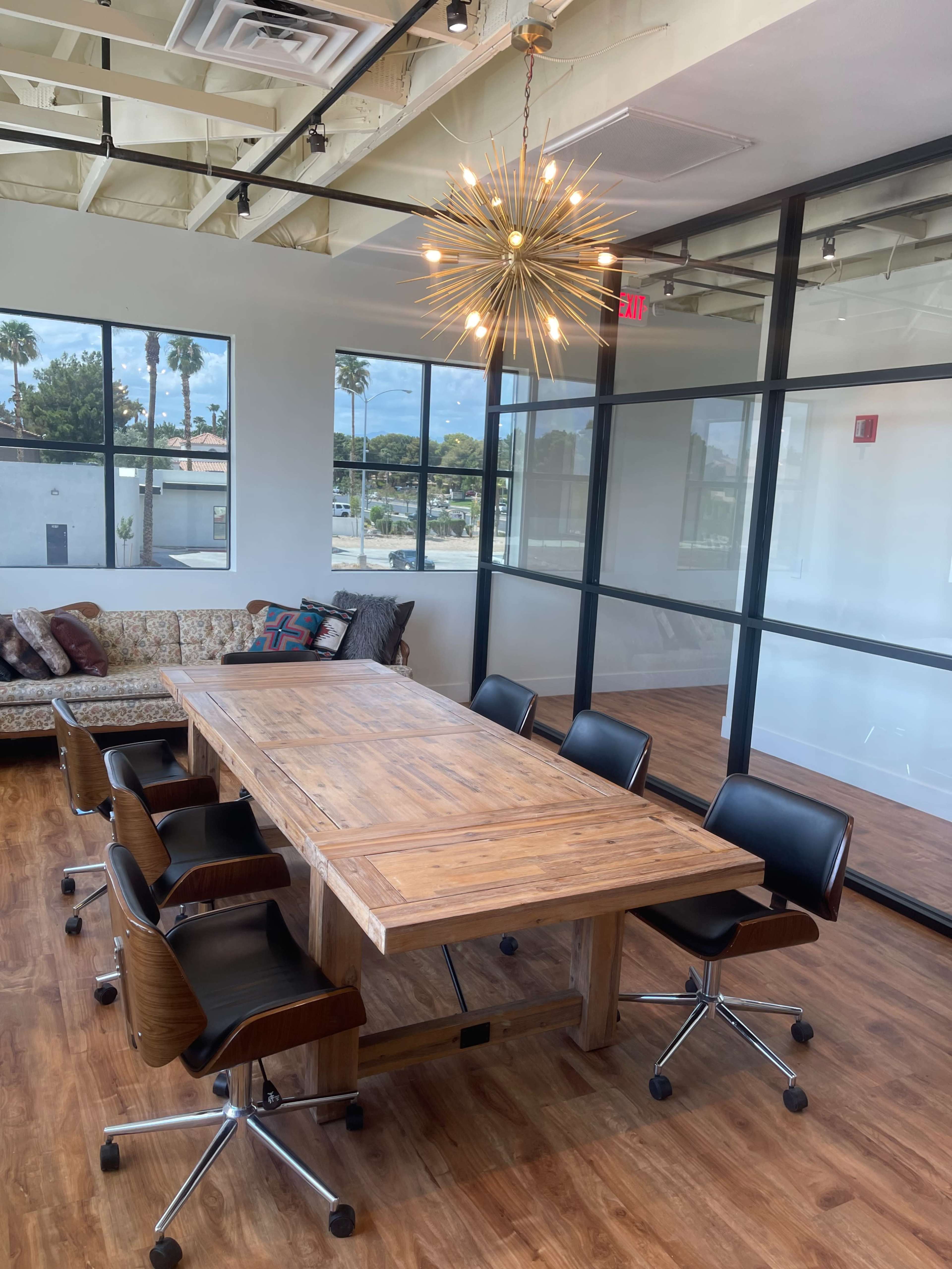 A modern conference room features a large wooden table surrounded by black swivel chairs, illuminated by a starburst chandelier, with large windows offering a view of palm trees outside.