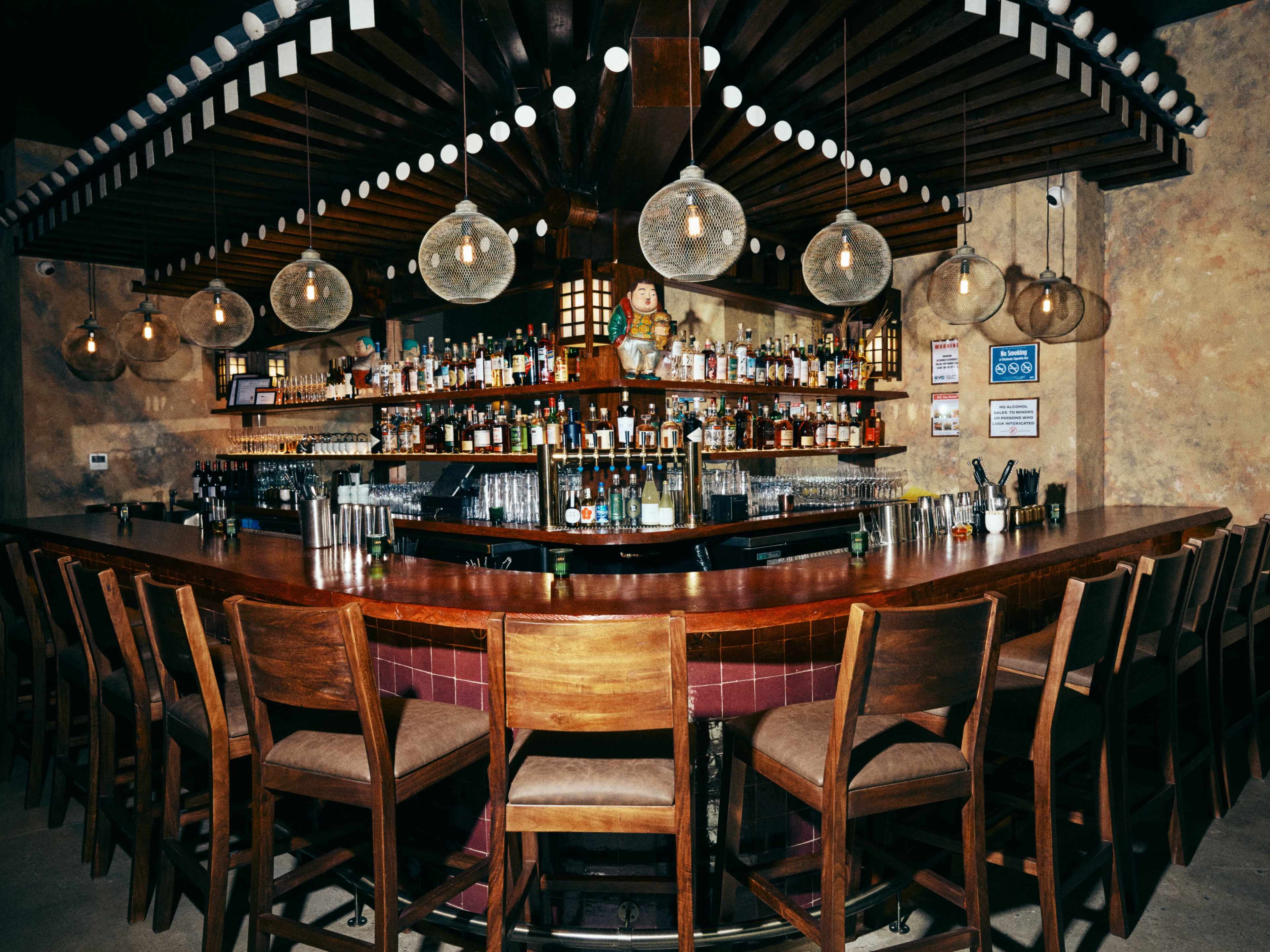 The image shows a curved wooden bar with high stools, surrounded by shelves filled with various bottles and decorative light fixtures overhead.