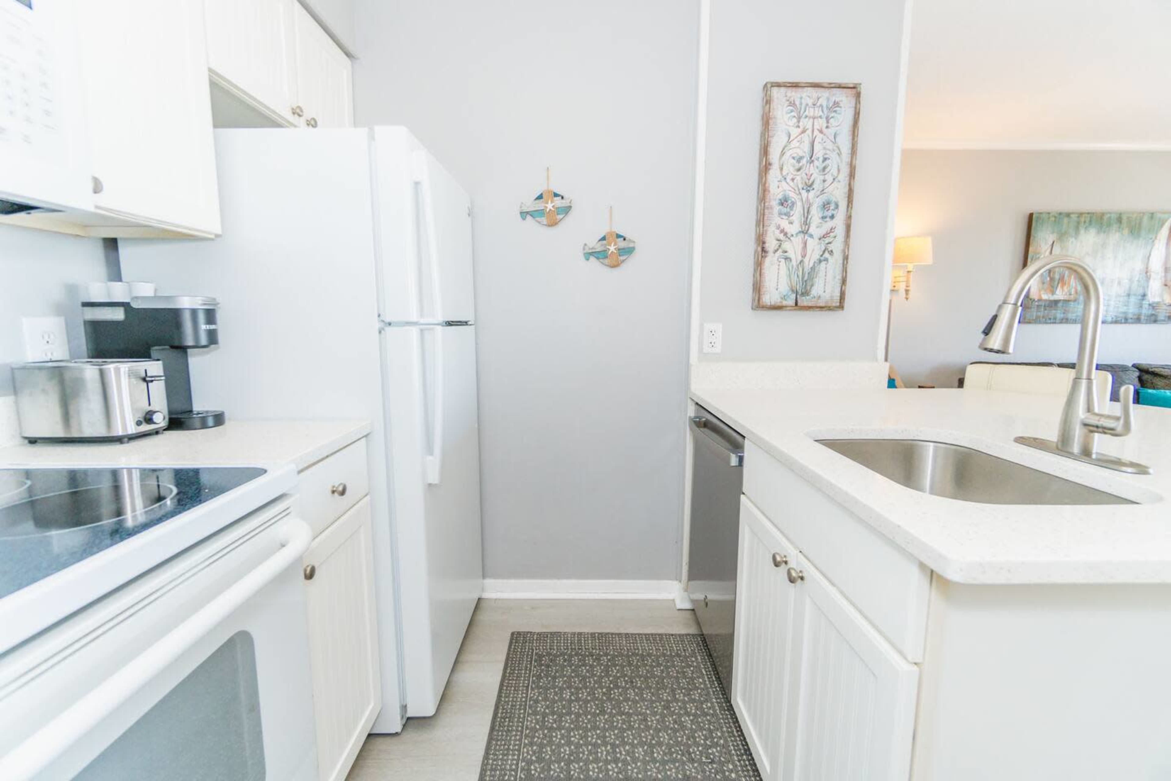 The image shows a small, modern kitchen featuring white cabinets, a stainless-steel sink, and appliances including a refrigerator and stove.