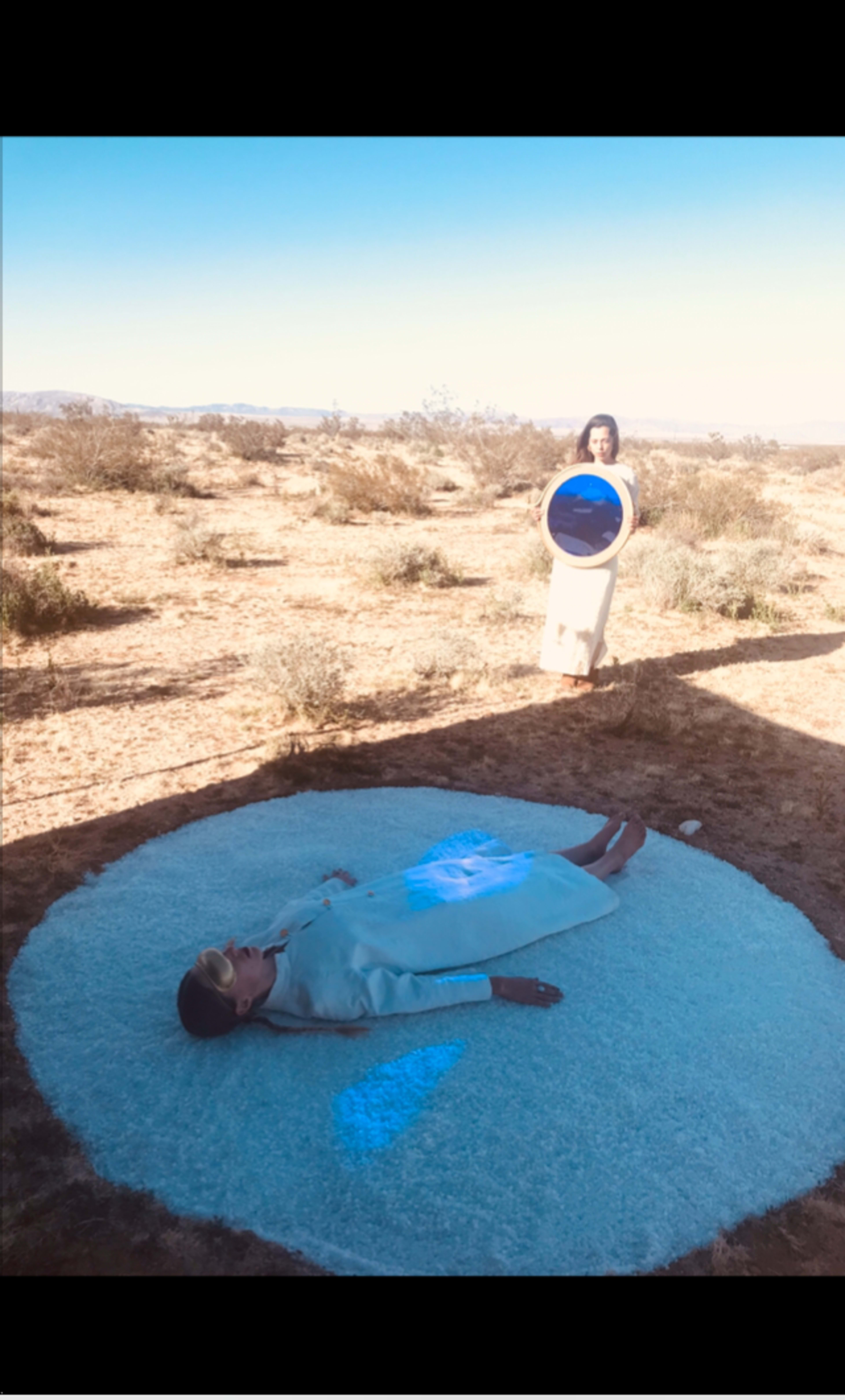 Two women are positioned in a desert landscape, one lying on a circular mound of white material while the other stands nearby holding a circular object that displays a blue image.