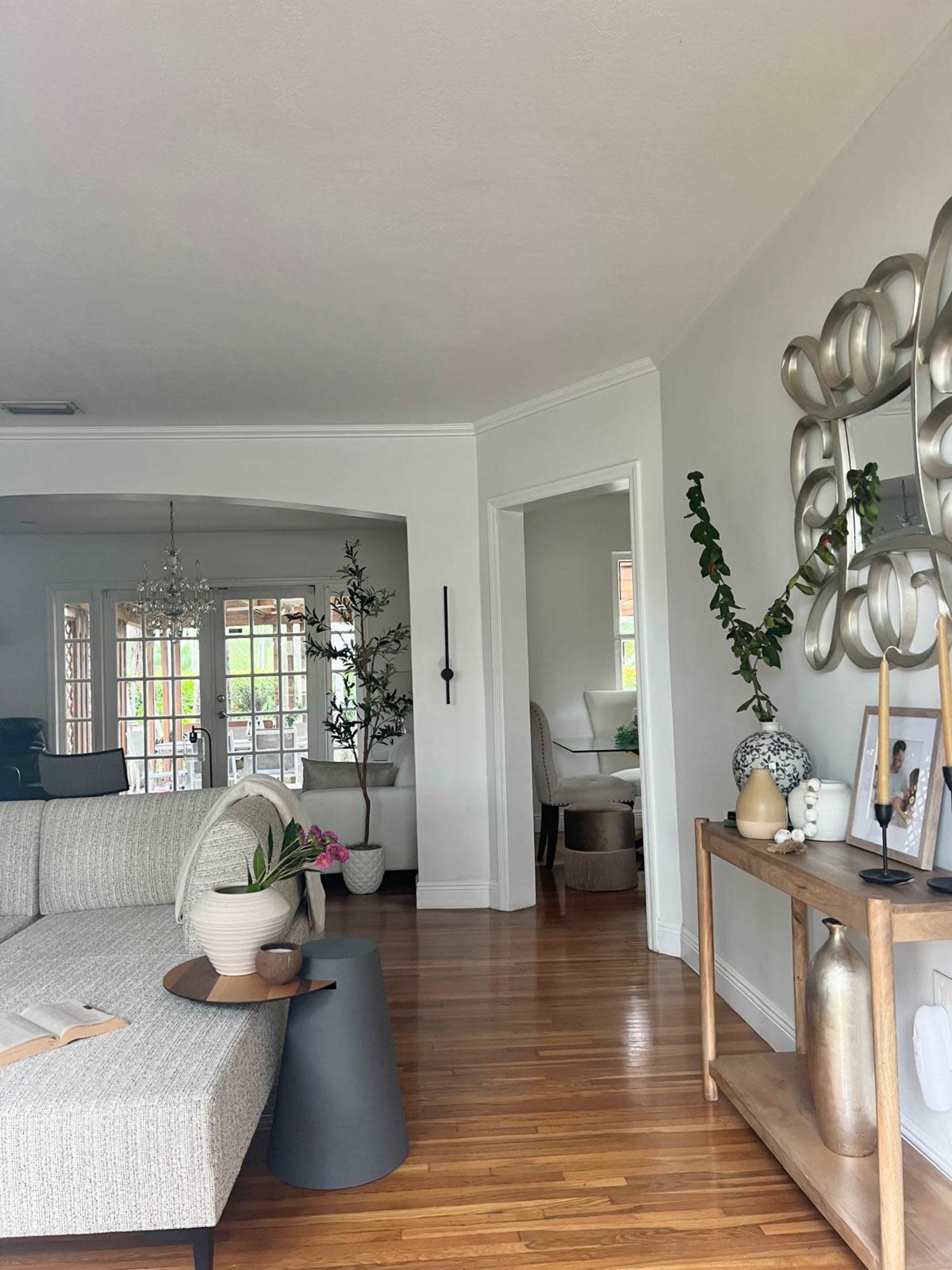 The image shows a stylish living space featuring a light-colored sofa, a wooden side table with decorative items, and a glimpse of a dining area through an open doorway.