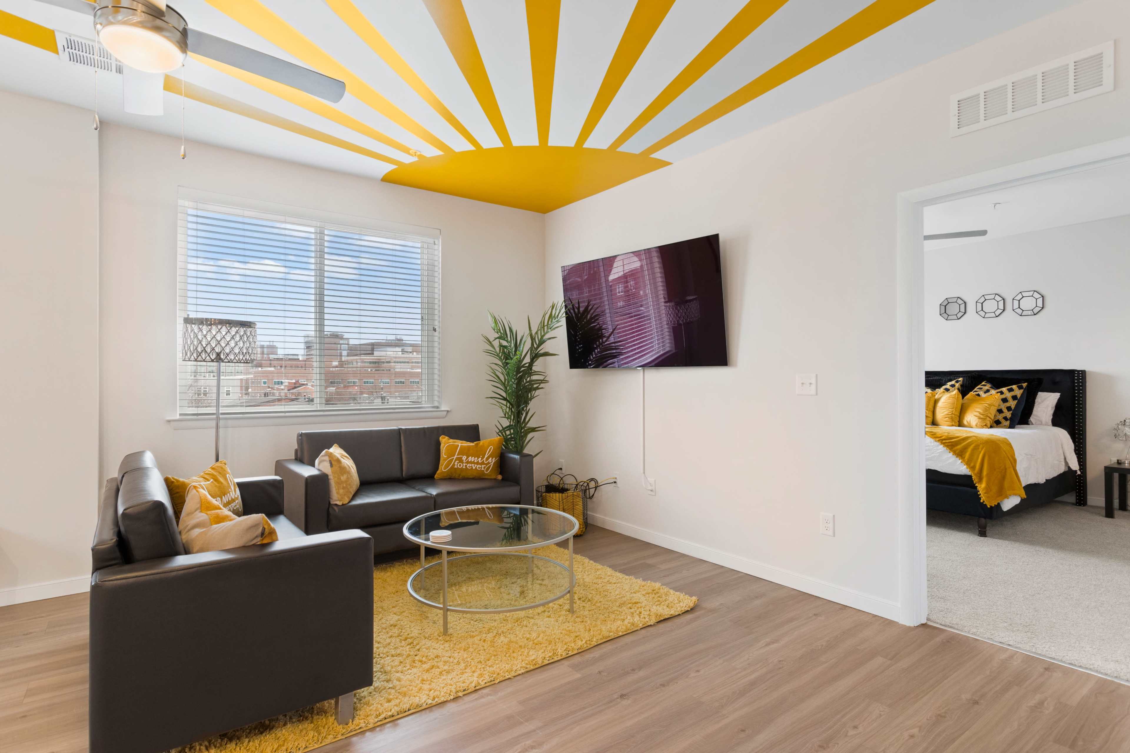 A modern living room features a black leather sofa set, a glass coffee table, and a yellow-themed ceiling with a sunburst design.