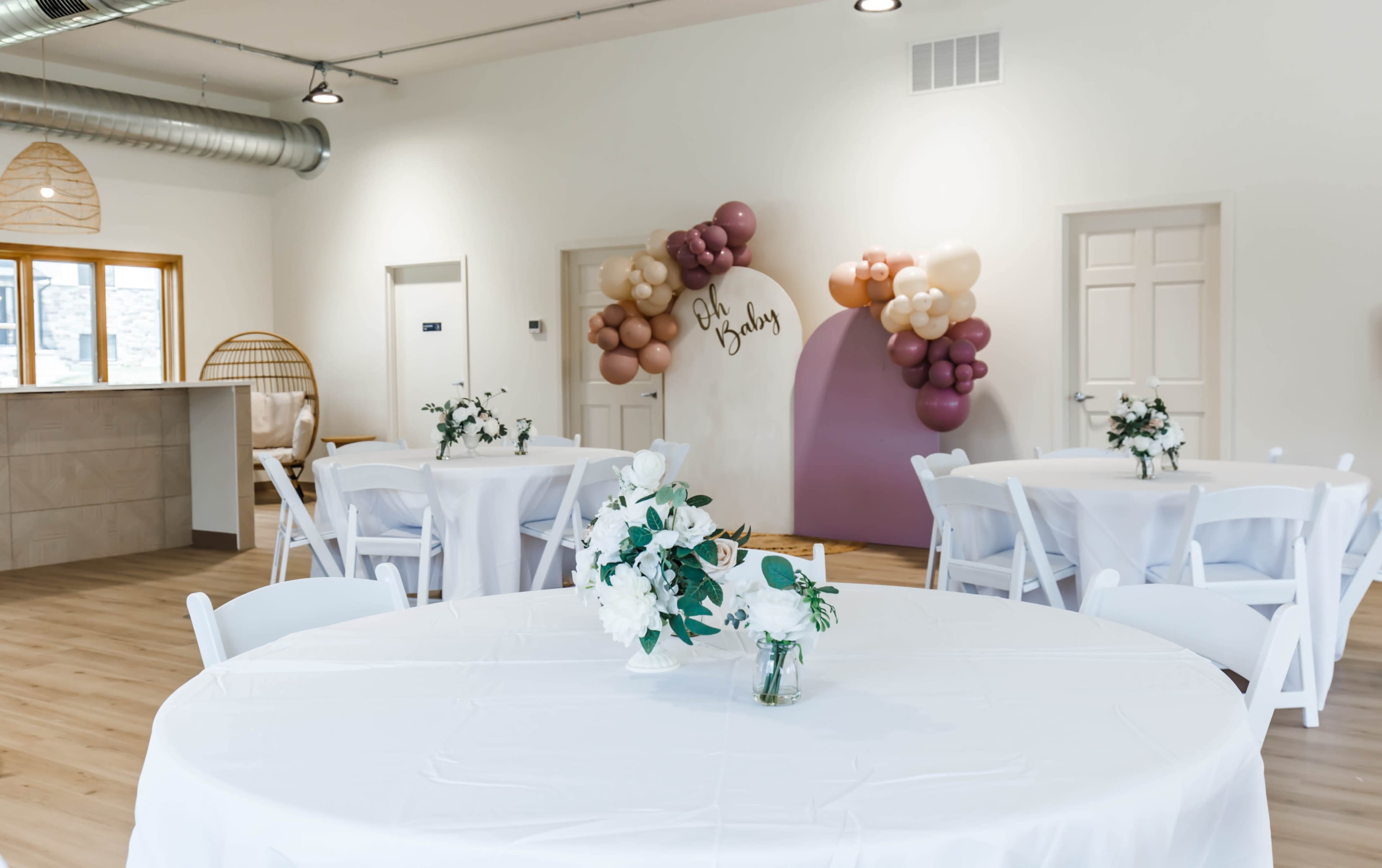 A bright event space decorated with white tables, floral centerpieces, and a balloon arrangement on the wall, featuring a sign that reads "Oh Baby."