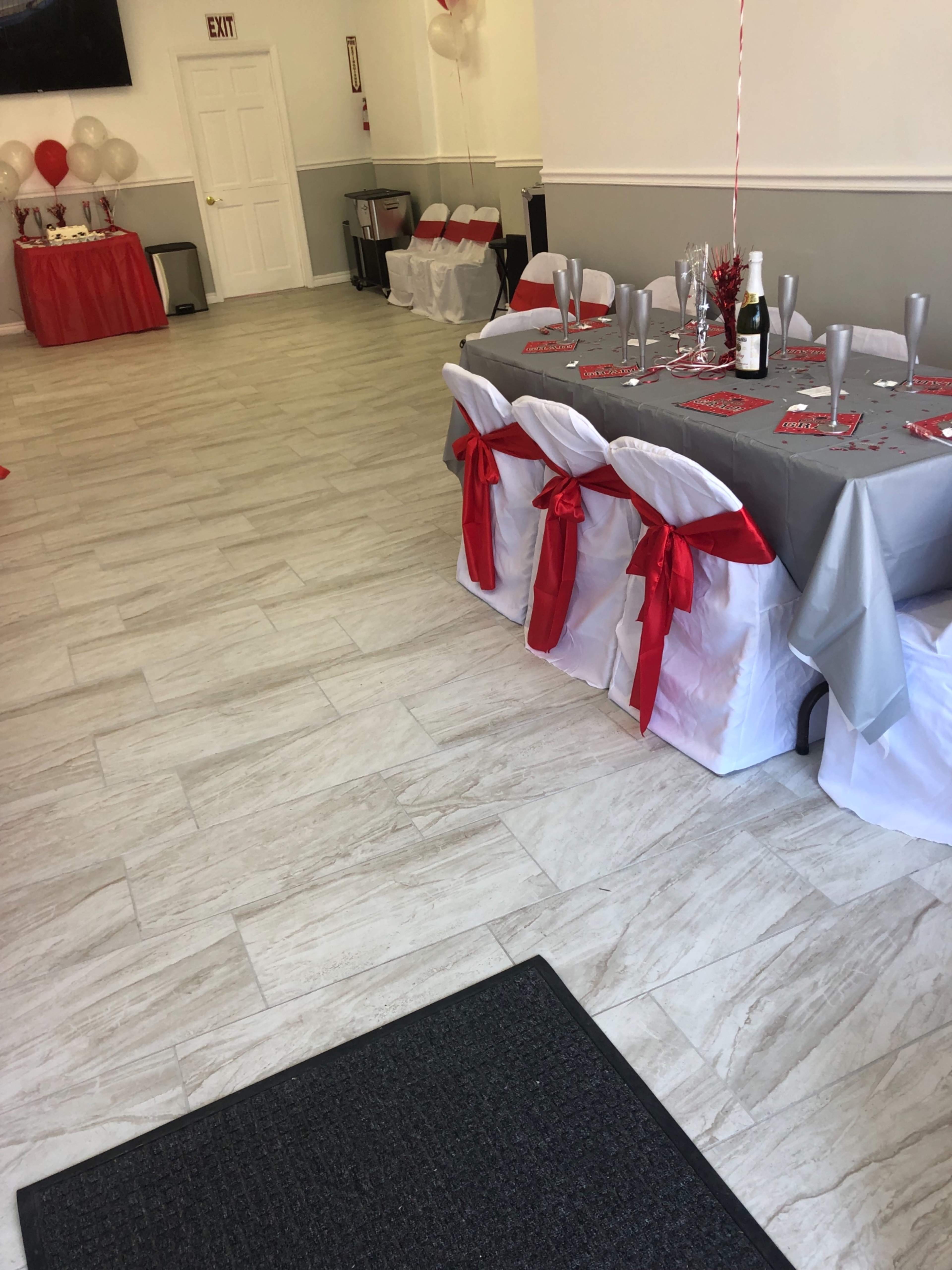 The image shows a dining area set for an event, featuring a table adorned with a gray tablecloth and white chairs tied with red ribbons, alongside a beverage station in the background.