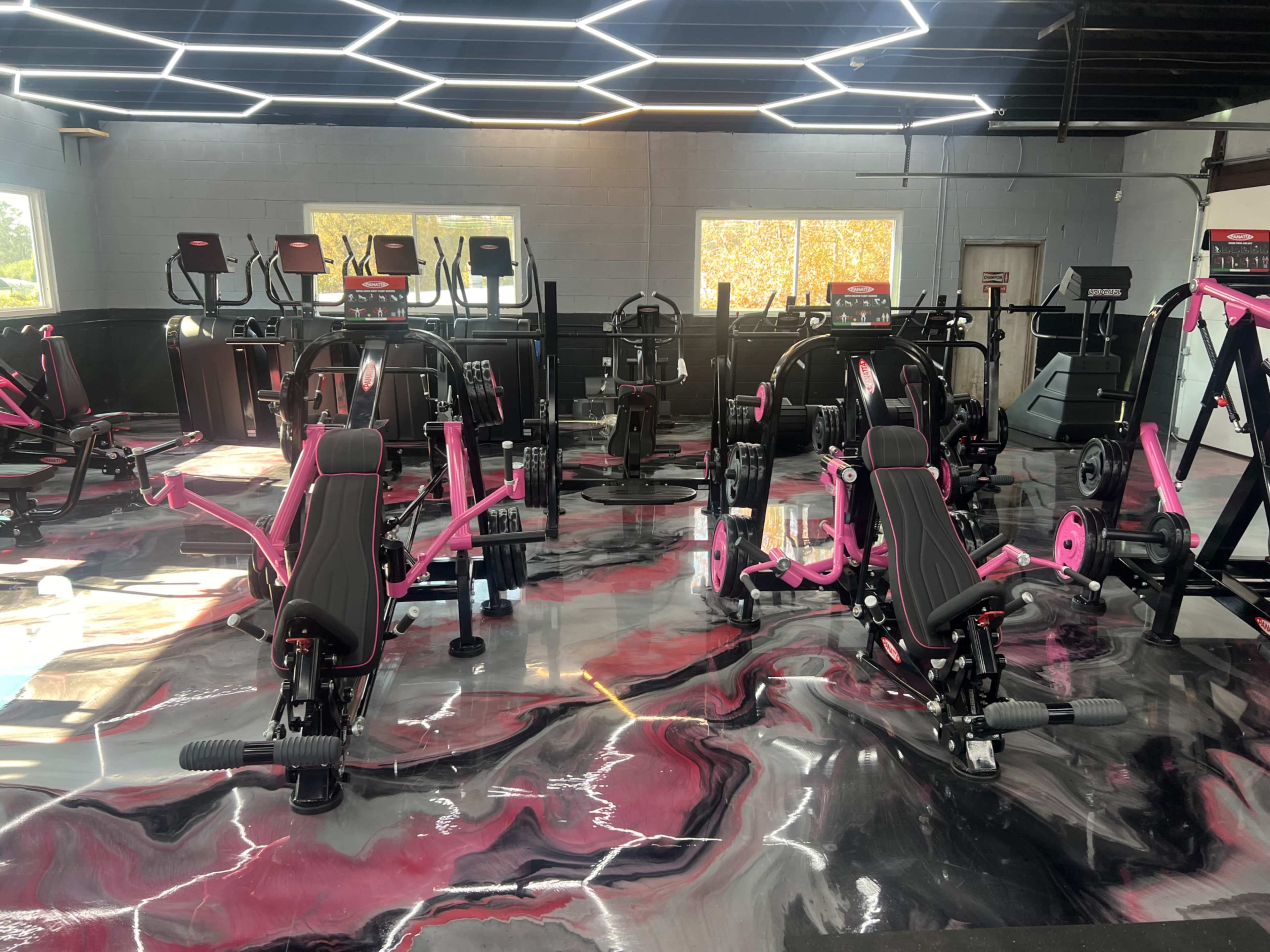 The image shows a modern gym interior featuring various exercise machines with black and pink designs on a glossy, marble-patterned floor.