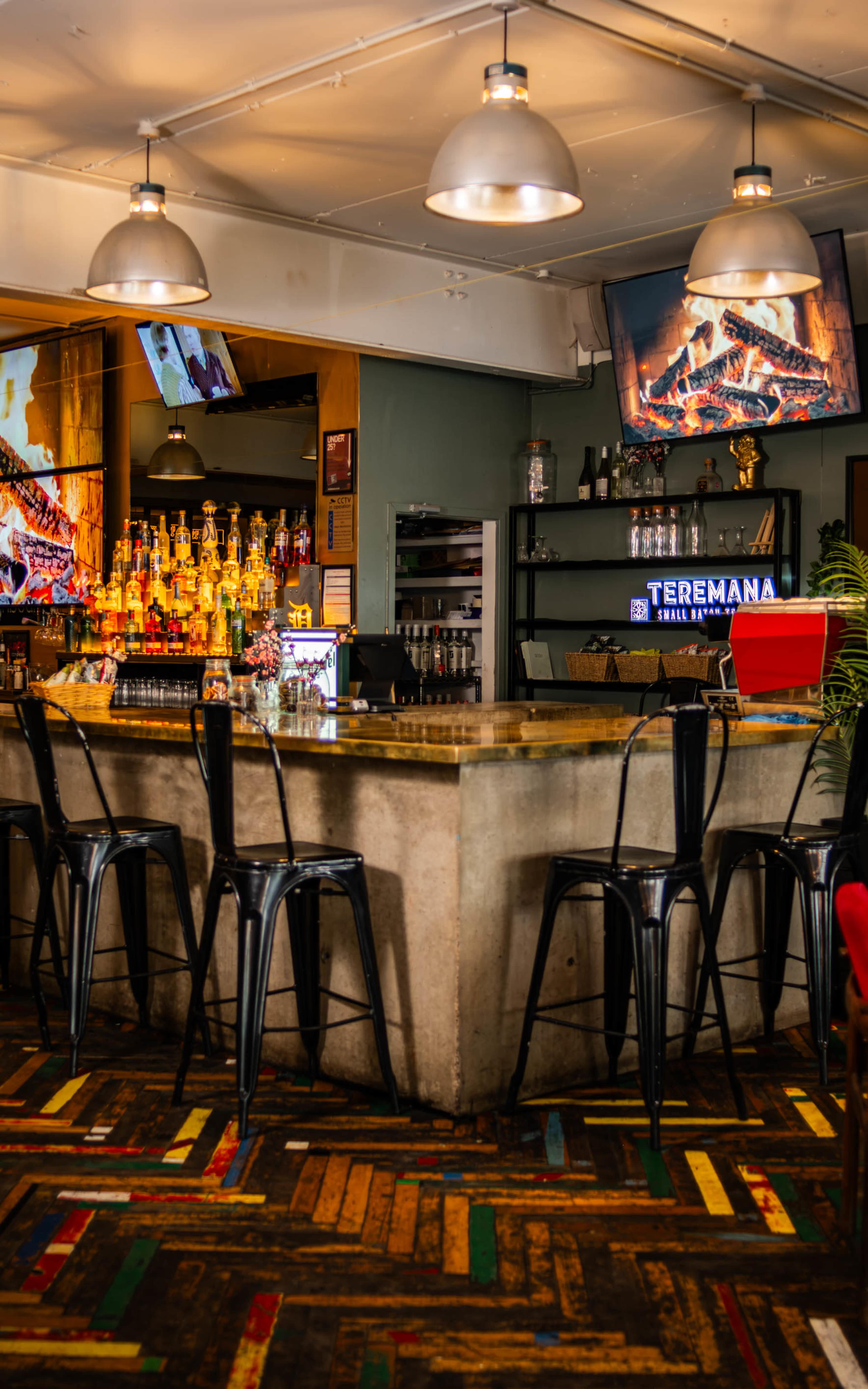 The image shows a bar area with a concrete counter, metal stools, a colorful patterned floor, and shelves stocked with various bottles in a lively setting.