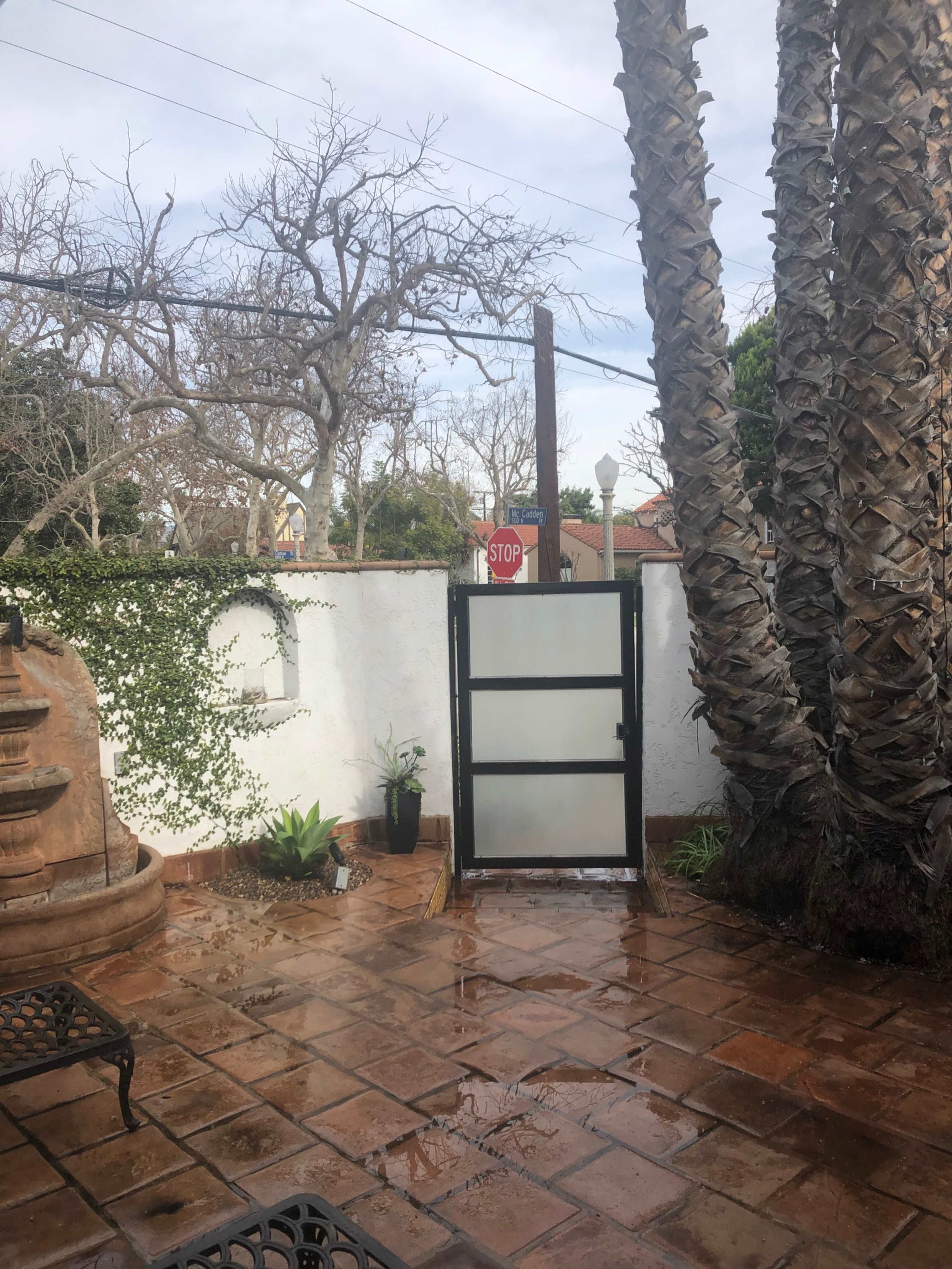 A courtyard with palm trees, a stop sign, and a partially open glass door, surrounded by a white wall and tiled floor.