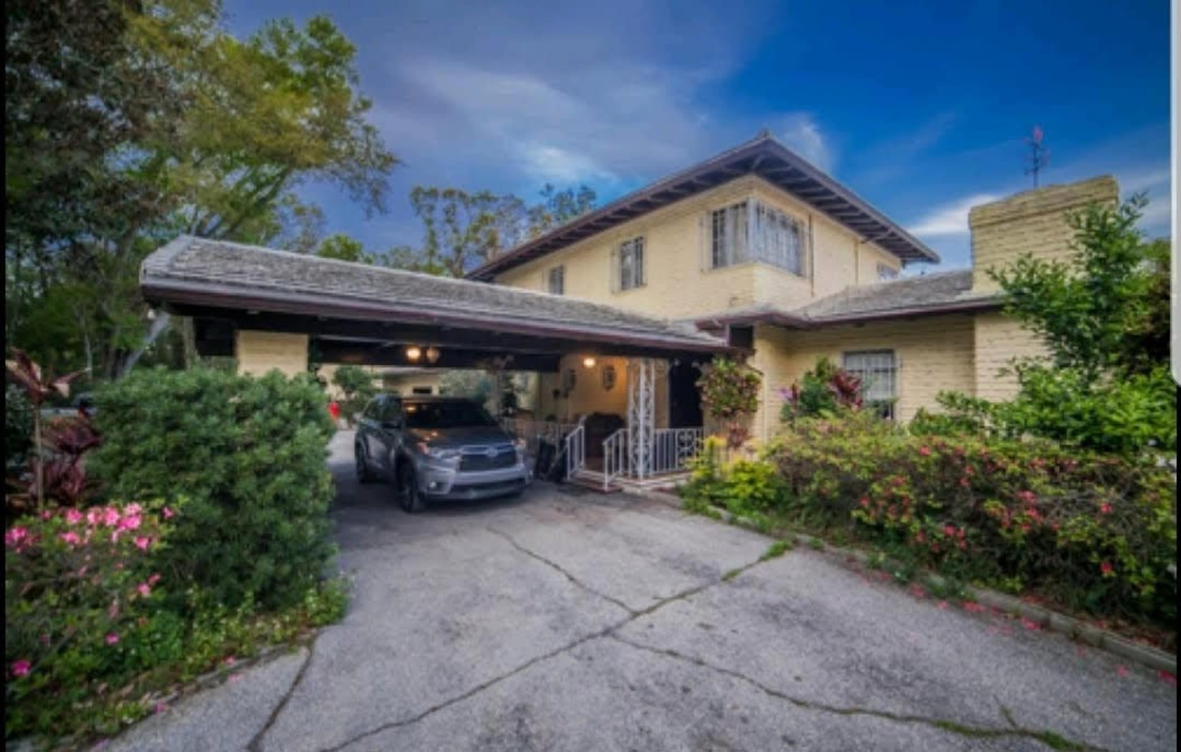 A two-story yellow house with a car parked in a driveway, surrounded by greenery and flowers.
