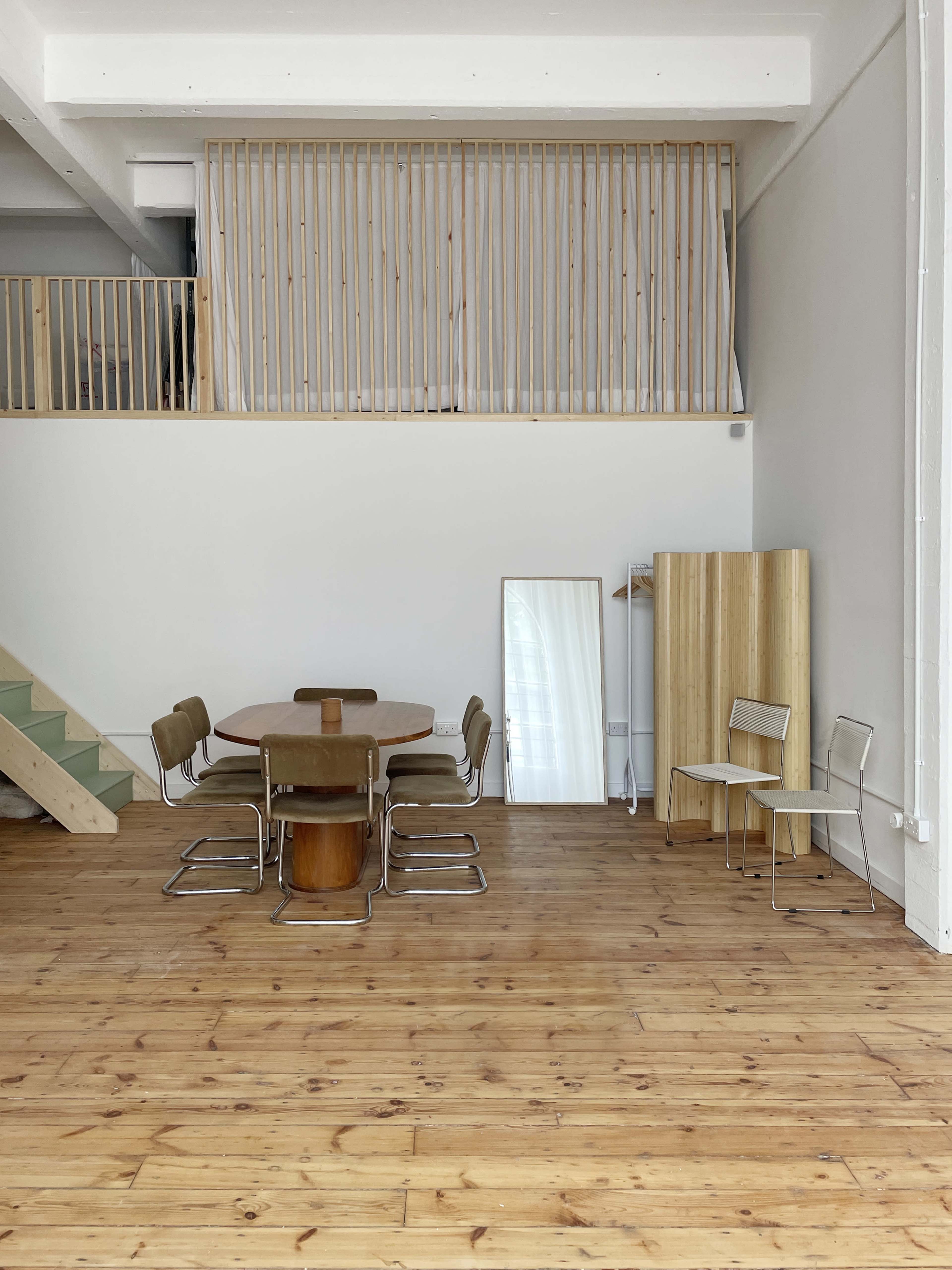 A minimalist interior space features a wooden dining table surrounded by metal chairs, with a staircase and wooden barriers in the background.
