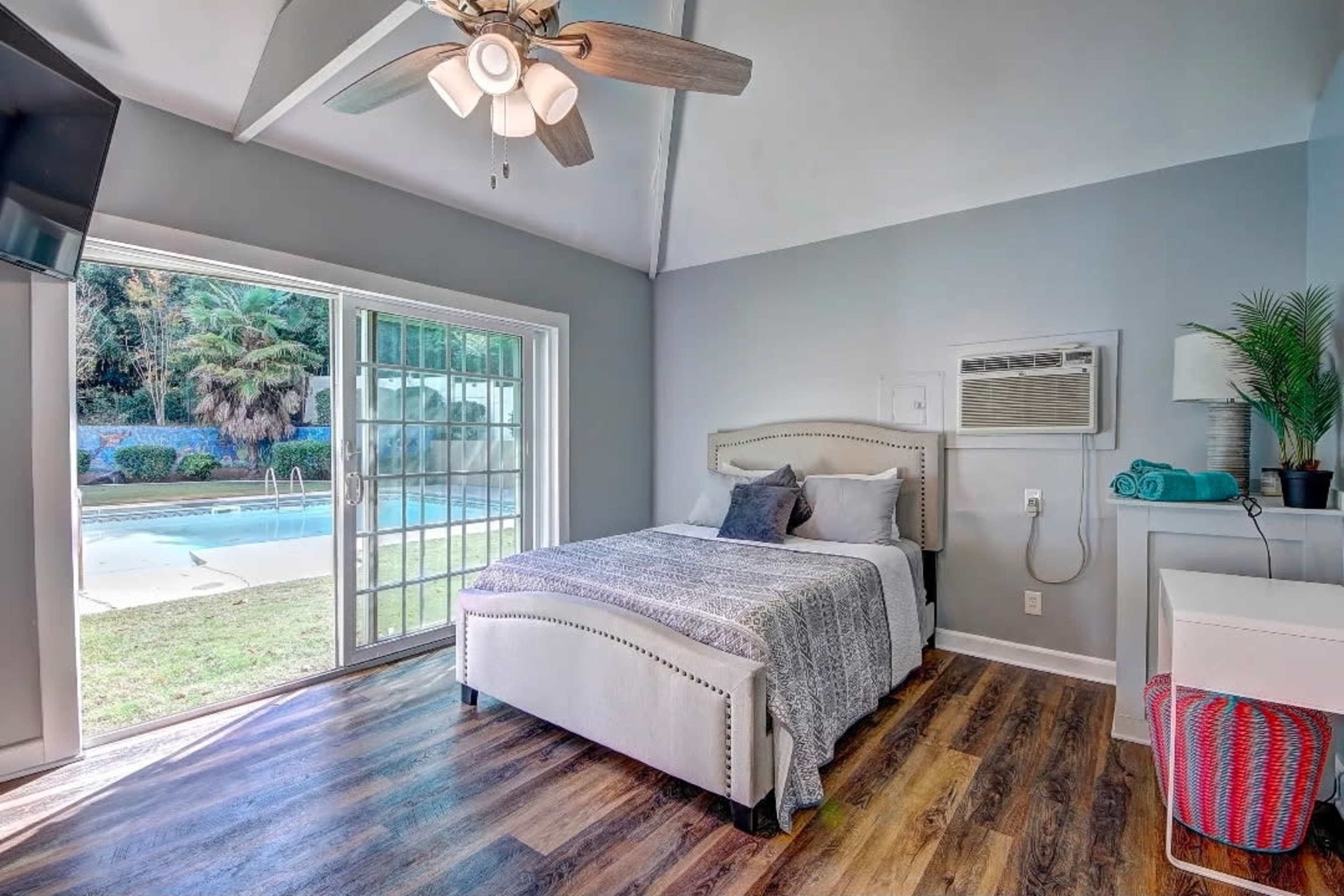 The image shows a brightly lit bedroom featuring a bed with gray linens, a ceiling fan, and large glass doors that open to a pool area outside.