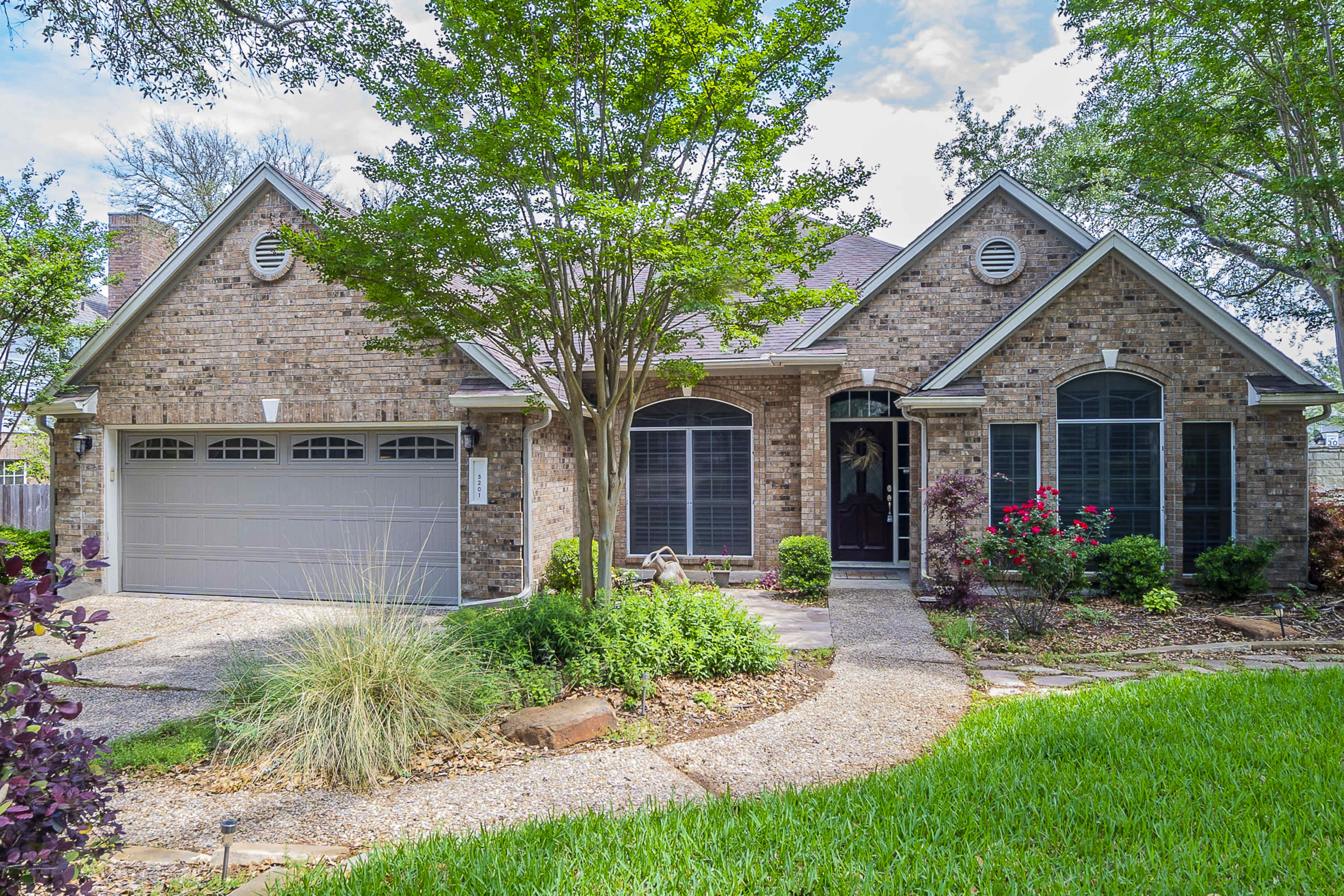 A brick house with a paved driveway, surrounded by landscaped gardens and shaded by trees.