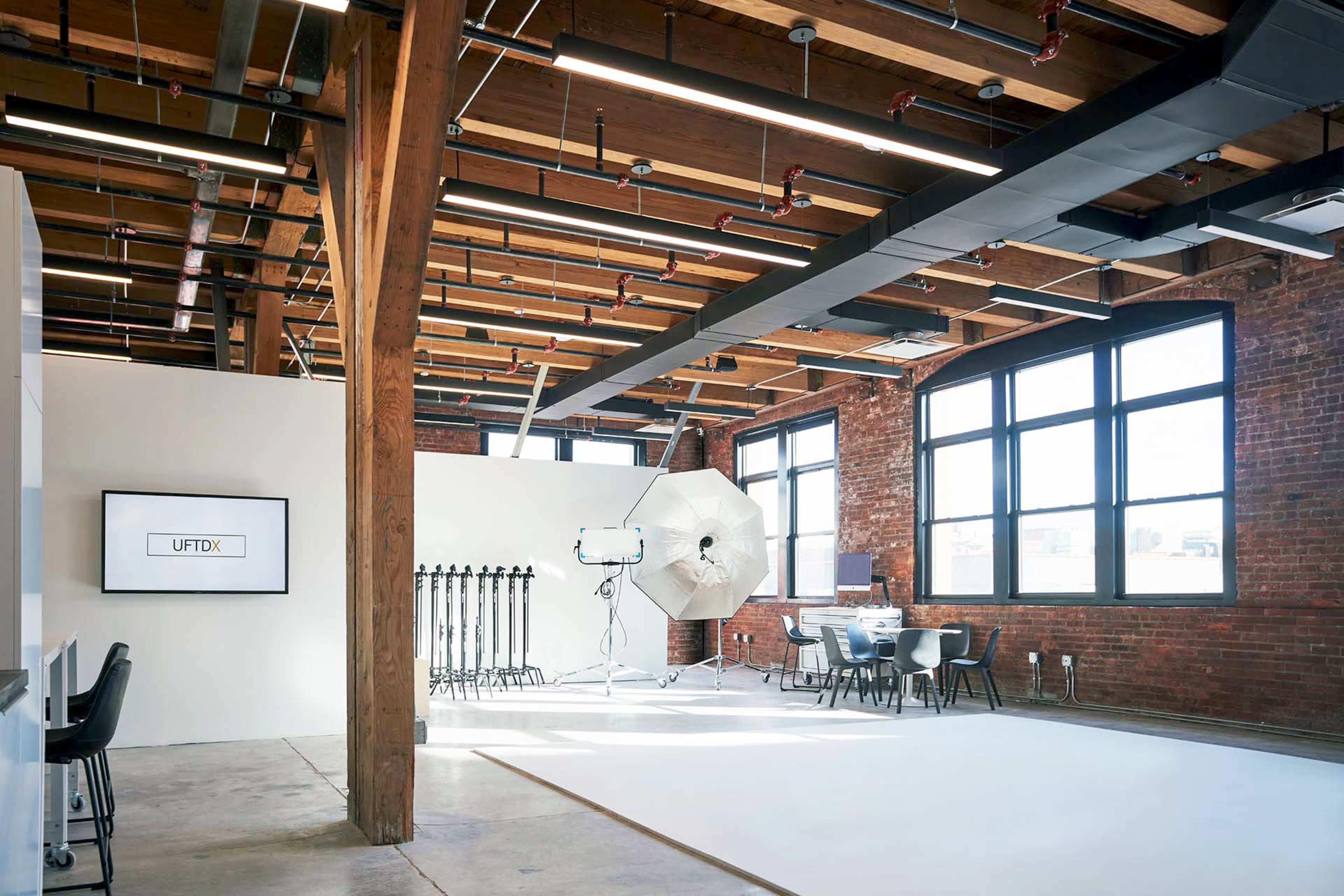 A spacious studio features exposed wooden beams, large windows, and photography equipment set up against a white wall.